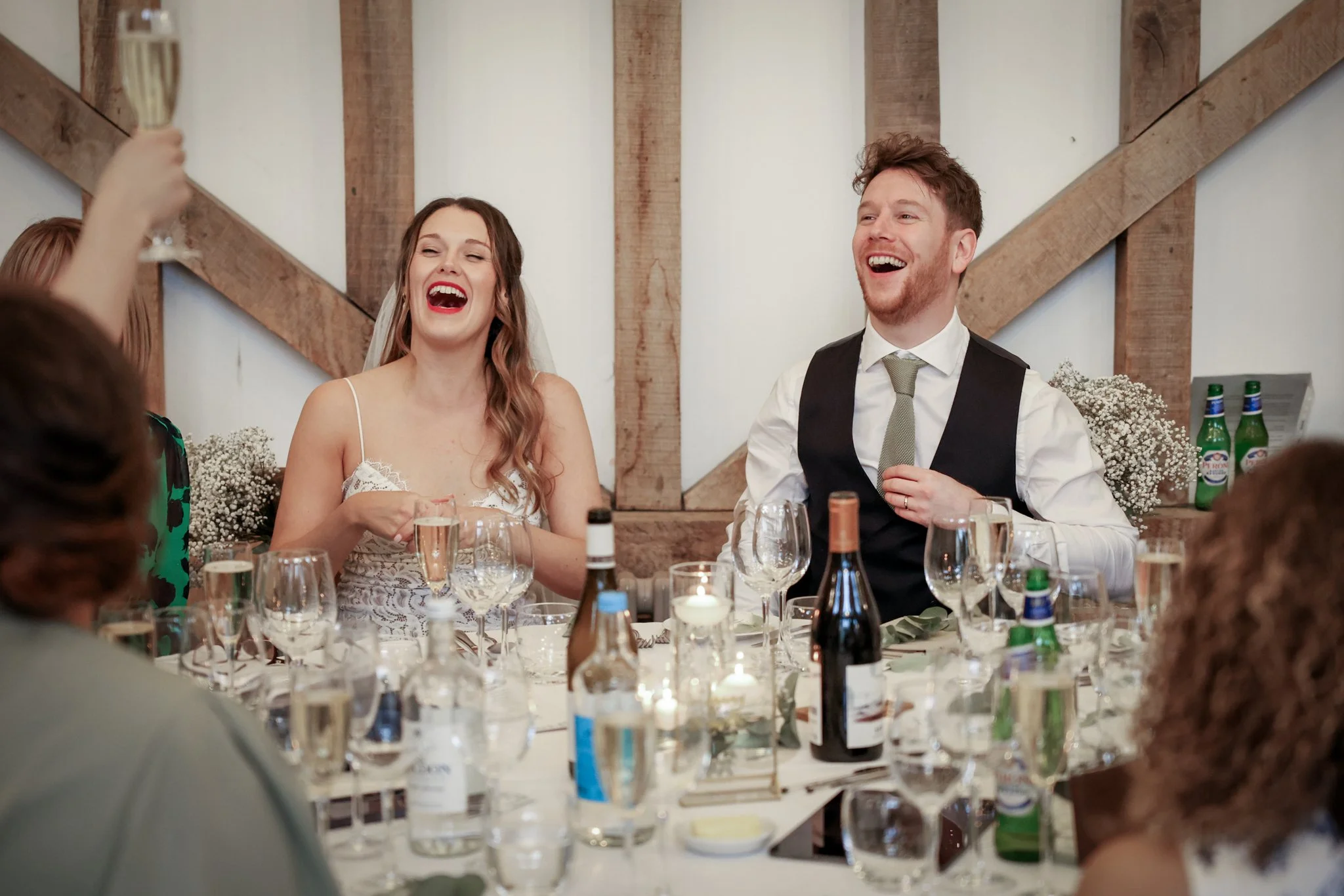 A bride and groom laughing at a wedding reception, seated at a table with drinks, candles, and floral decorations, in a rustic indoor setting.