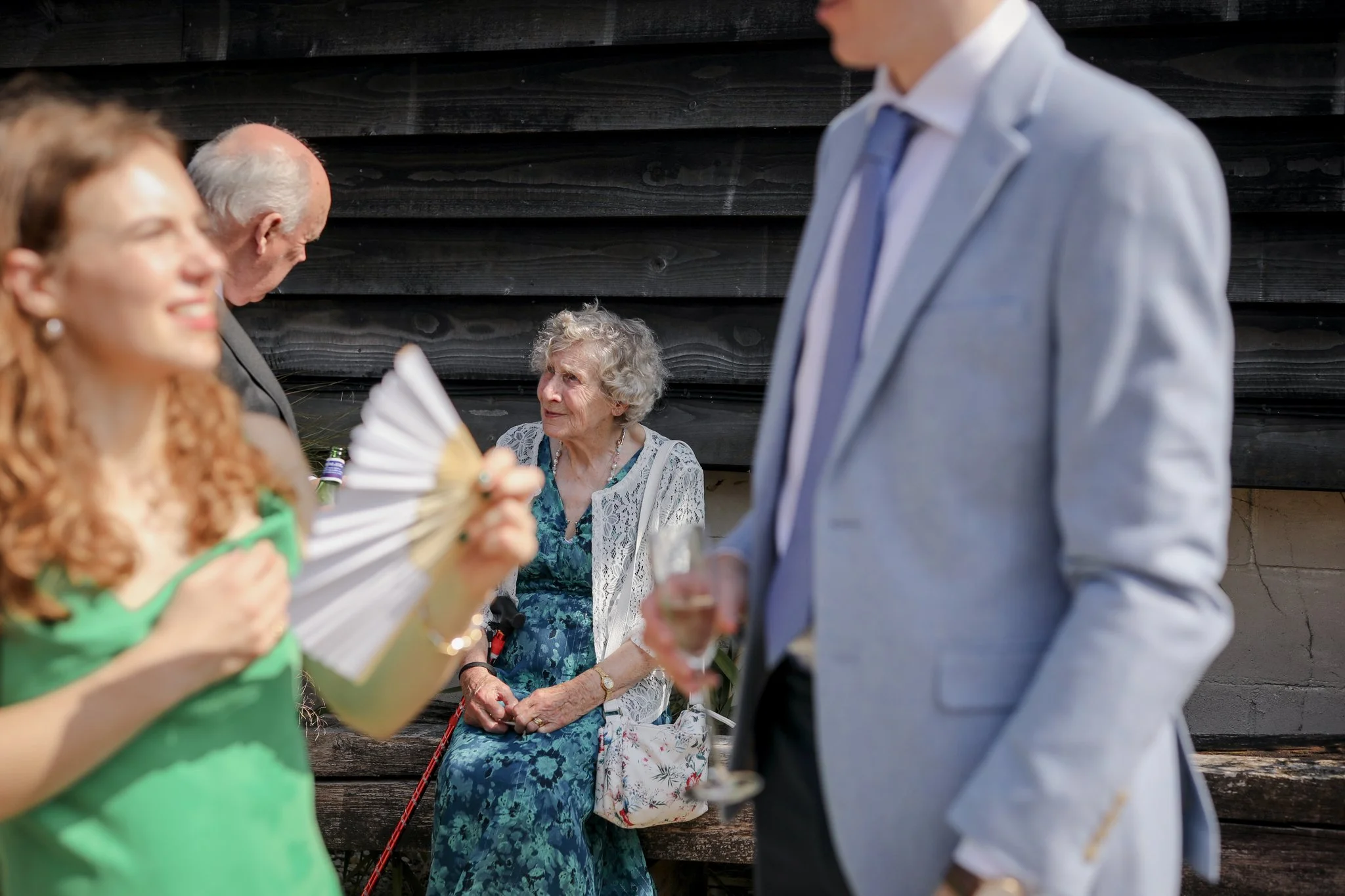 Blurred woman with red hair wearing a green dress holding a fan on the left side of the image. Elderly woman with curly gray hair wearing a blue dress with a white lace cardigan sitting on a bench in the background. Partially visible man in a gray su