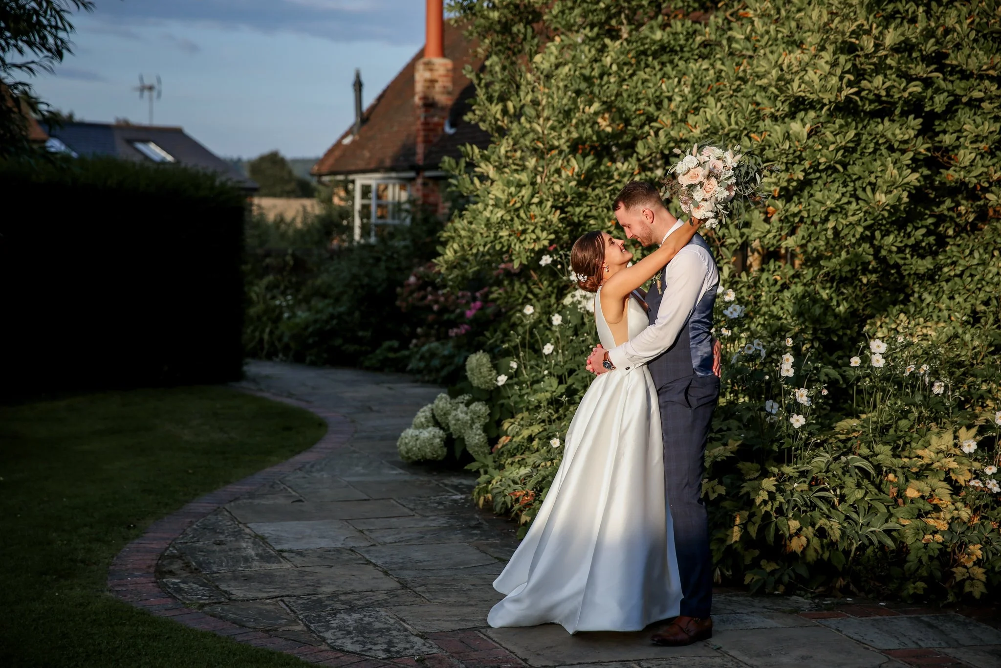 A bride and groom are embracing outdoors on a garden path, surrounded by lush green bushes and flowers. The bride wears a white wedding gown and the groom wears a dark suit. They are looking into each other's eyes, with the bride holding a bouquet of