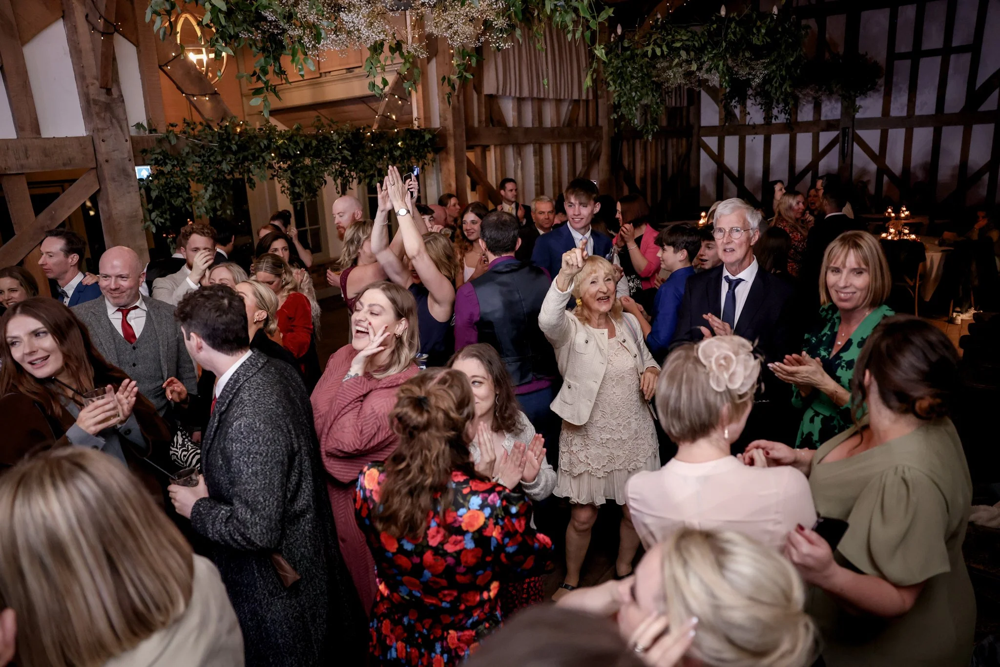 A group of people at an indoor celebration or party, dressed in formal and semi-formal attire, dancing and smiling. The venue has rustic wooden walls and ceiling with decorative greenery and string lights hanging from above.
