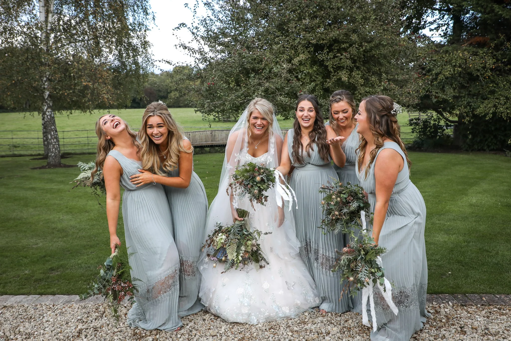 A bride in a wedding dress holding a bouquet, surrounded by five bridesmaids in matching light blue dresses, all smiling and laughing outdoors in a green park with trees in the background.