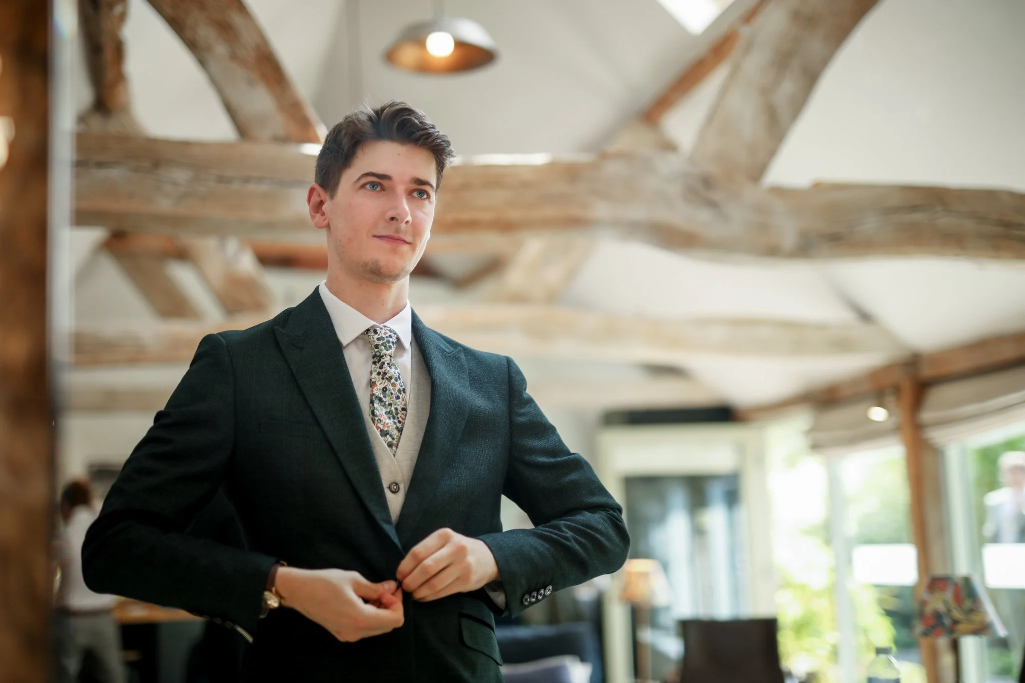 A young man in a suit standing indoors, with wooden beams and windows in the background.