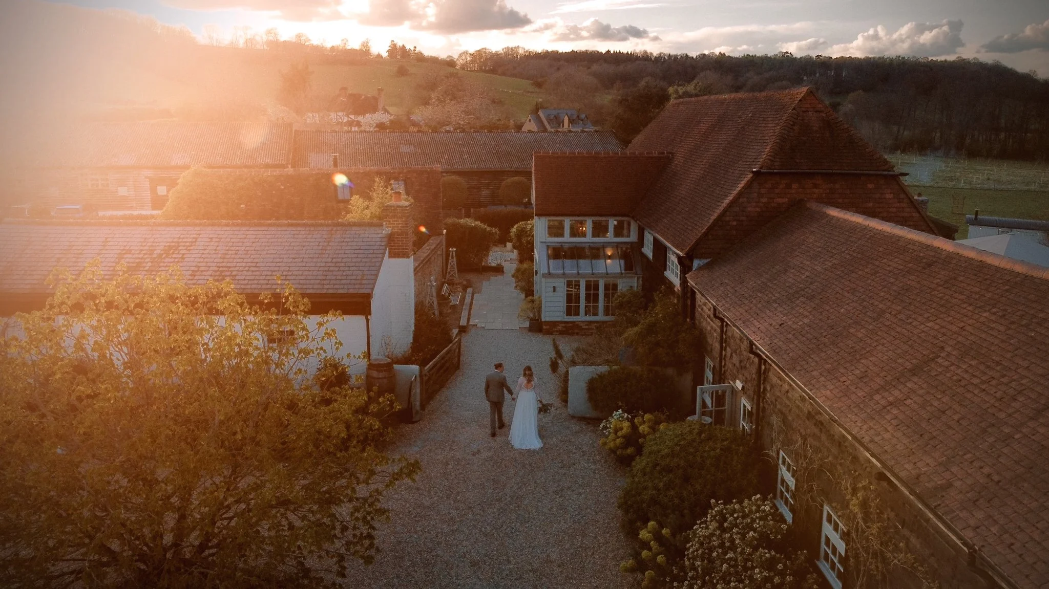 A couple walks hand in hand outside a rustic brick and wood house at sunset, with rolling hills and a cloudy sky in the background.