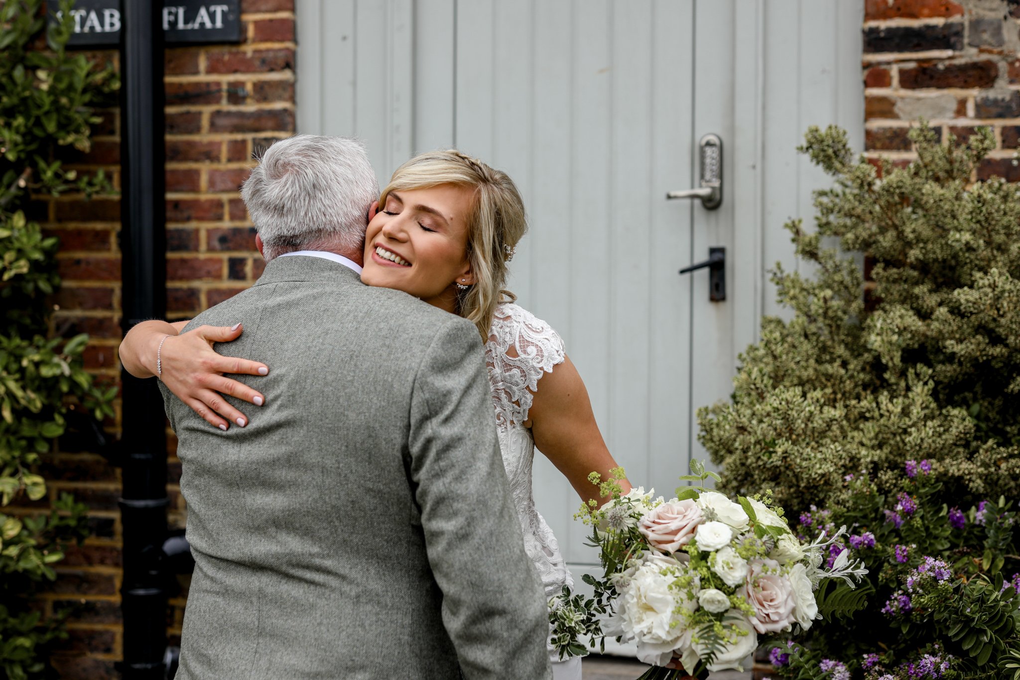 A woman in a wedding dress hugging an older man in a gray suit outside near a brick wall and a light blue door, holding a large bouquet of flowers.
