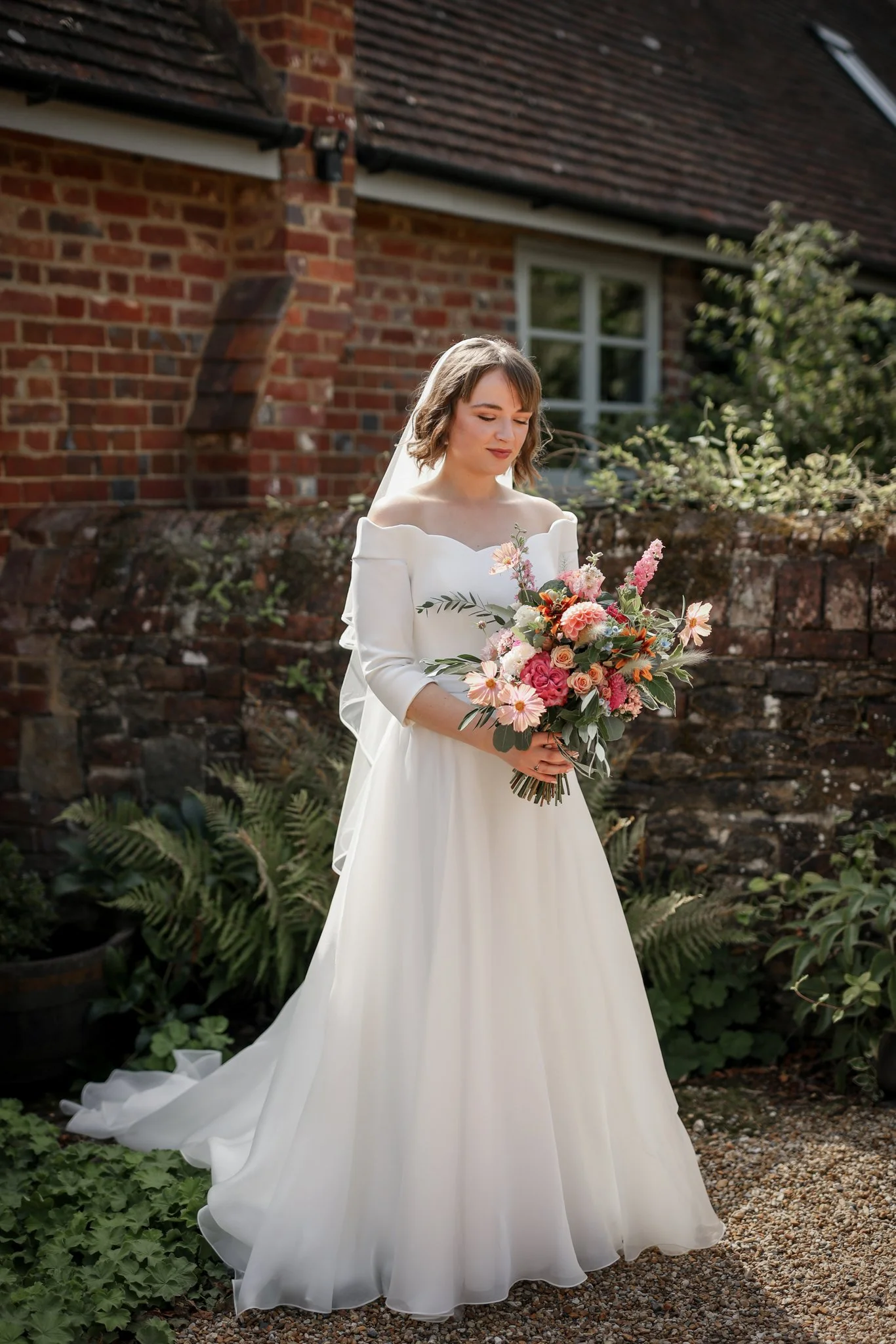 A bride in a white off-shoulder wedding dress with ruffled sleeves holding a large bouquet of pink, white, and peach flowers with greenery, standing outdoors near a brick house with a window and lush green plants.