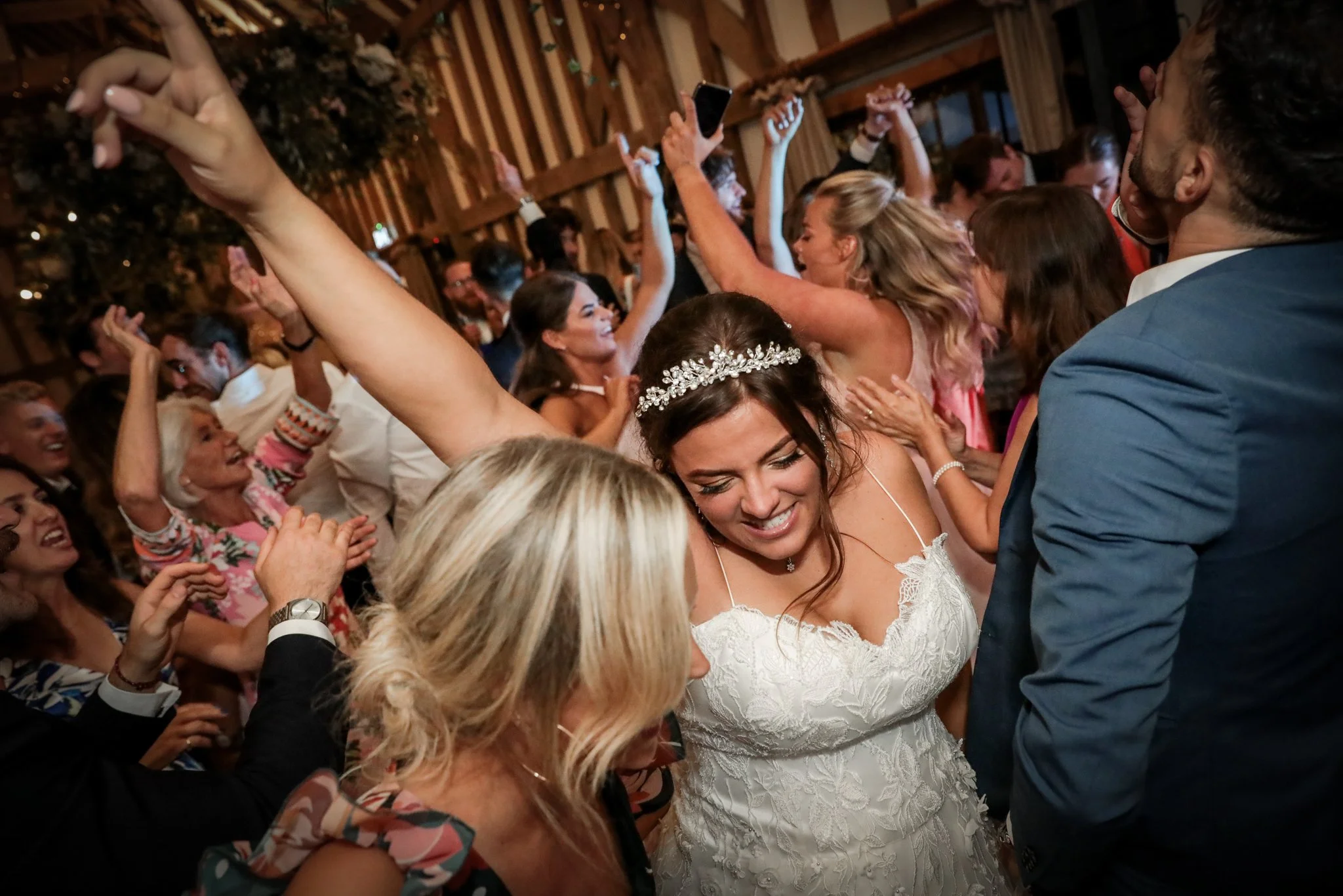 A group of people dancing and celebrating at a wedding in a rustic barn with wooden beams. The bride, wearing a white lace gown and a decorative headband, is smiling. Guests are raising their hands and enjoying the moment.