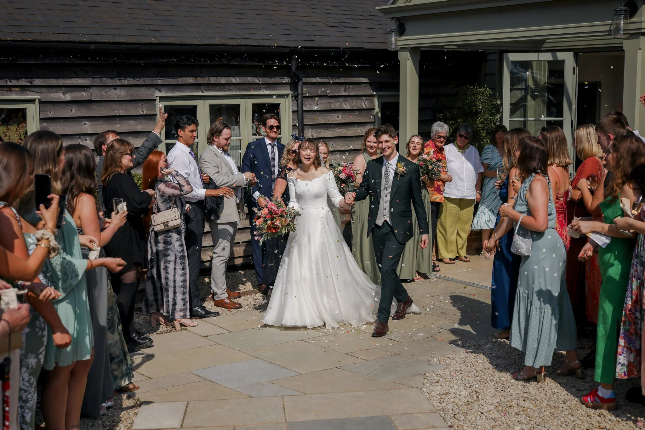A wedding celebration outdoors with a bride and groom walking hand in hand, surrounded by guests. Guests are throwing confetti and smiling. The setting includes a rustic wooden building in the background.
