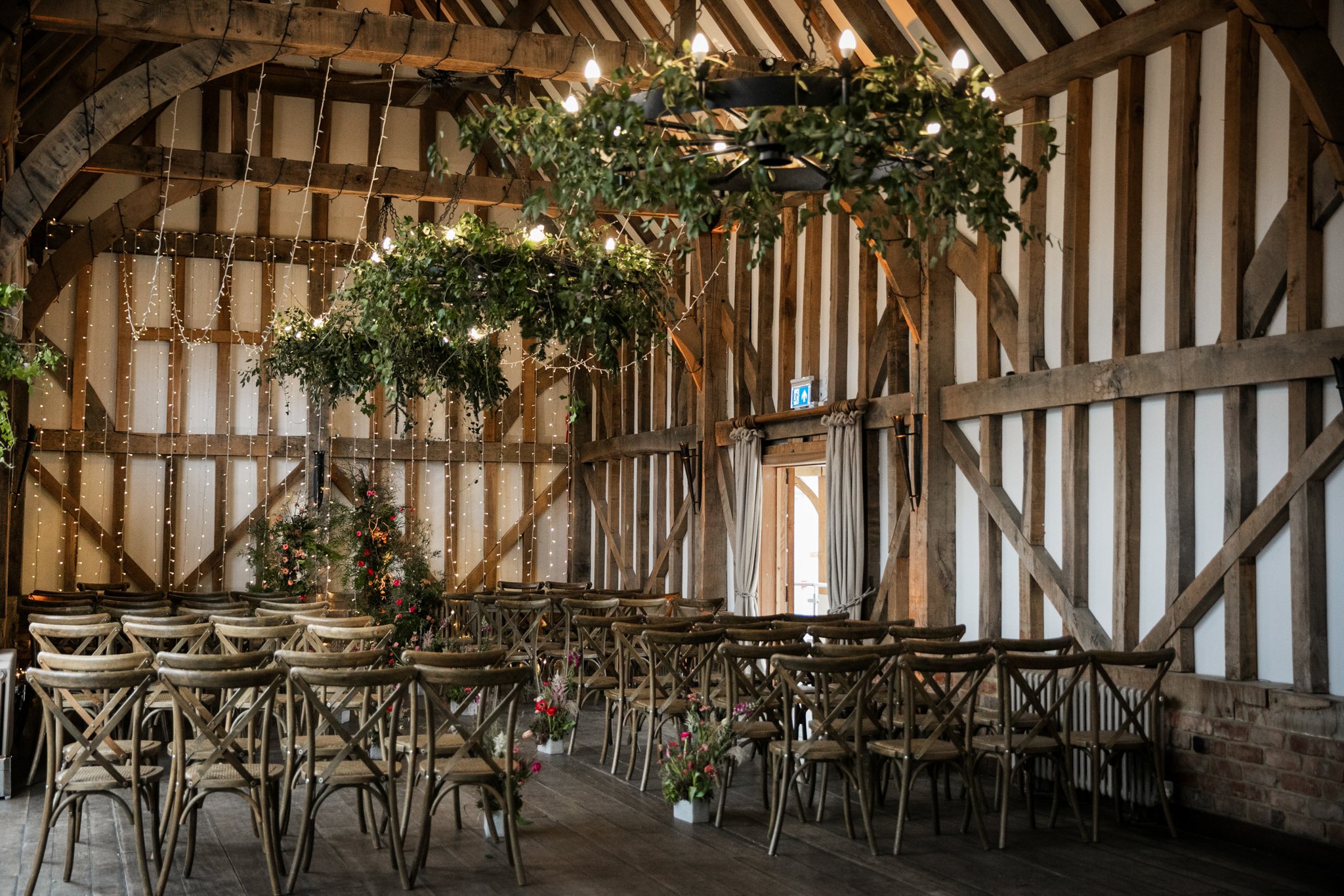 Interior of a rustic barn decorated for an event with rows of wooden chairs, hanging greenery with lights, and twinkling string lights on the walls.
