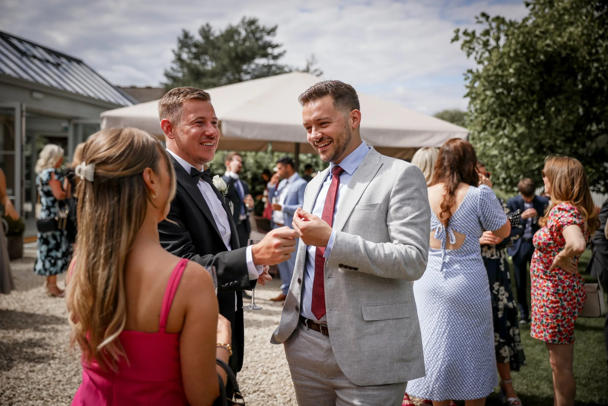 Group of people at an outdoor celebration, with two men in suits smiling and talking, while others mingle in the background under a white canopy.