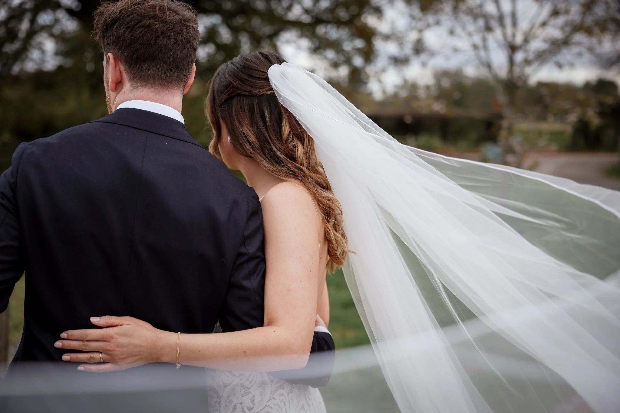 A bride and groom embrace outdoors, the bride wearing a wedding dress and veil, and the groom in a dark suit.