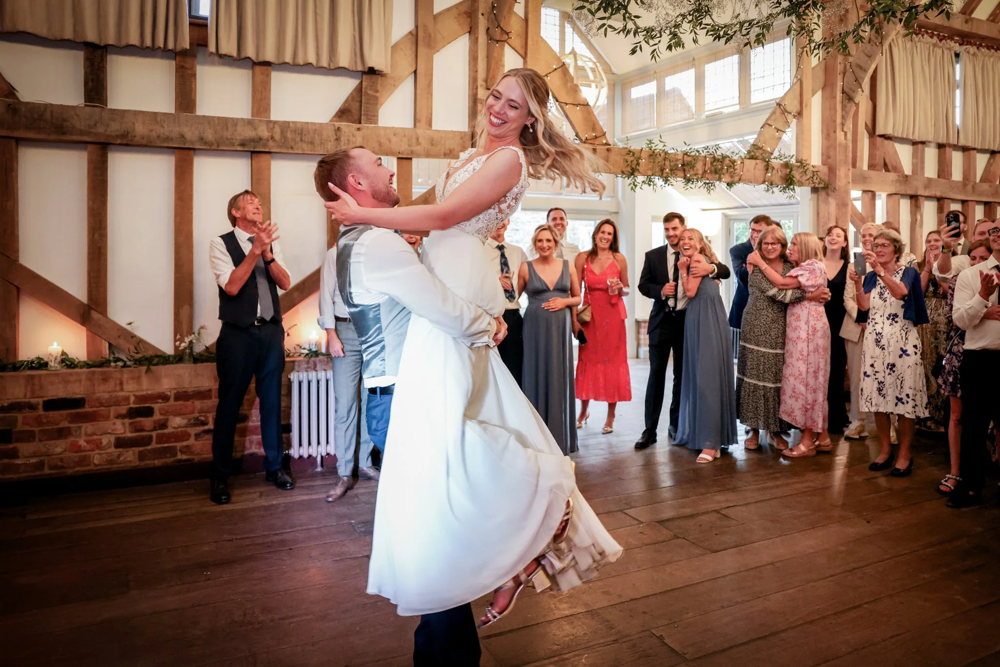 A bride and groom are dancing at their wedding reception, surrounded by cheering guests inside a rustic decorated venue with wooden beams and string lights.