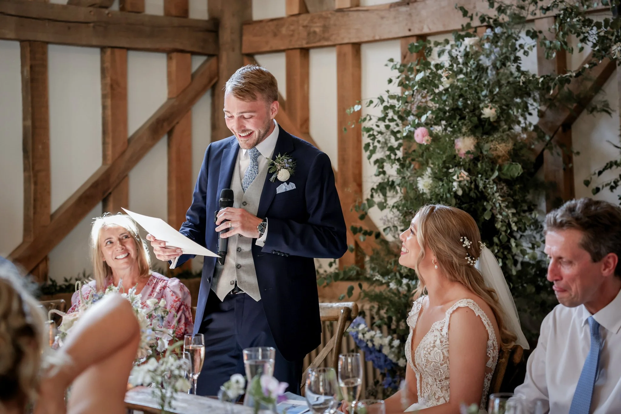 A man in a suit giving a speech with a microphone at a wedding reception. Women at the table are listening and smiling, with floral arrangements and glasses of champagne on the table. The setting is rustic with wooden beams and greenery.