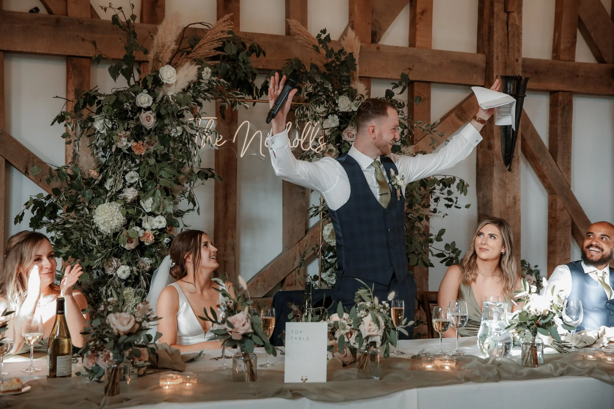 A wedding reception with a groom giving a speech, surrounded by bridesmaids and groomsmen, and decorated with floral arrangements and greenery.