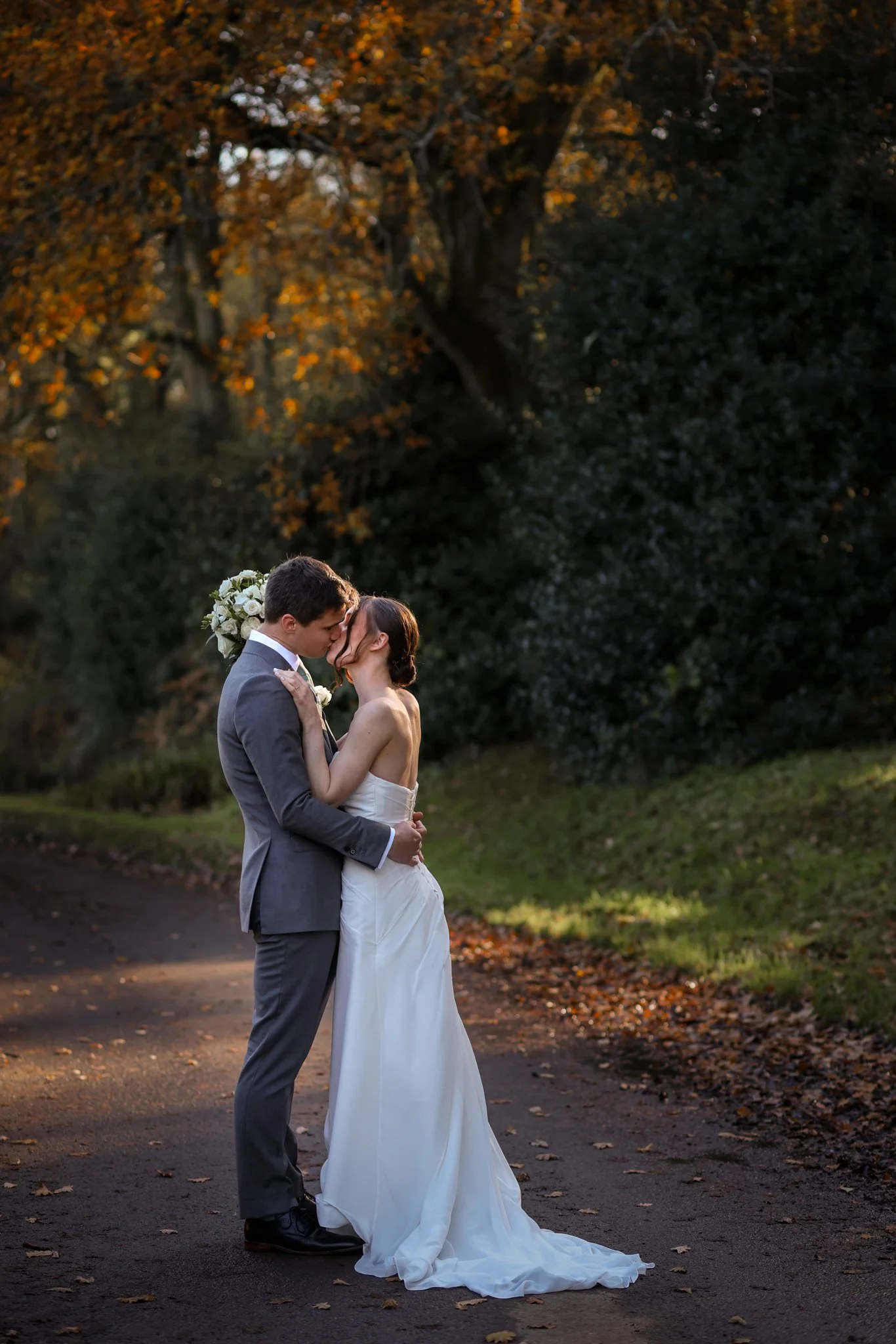 A bride and groom in a loving embrace during their wedding in an outdoor setting with trees and fallen leaves, with the groom holding a bouquet of white flowers.