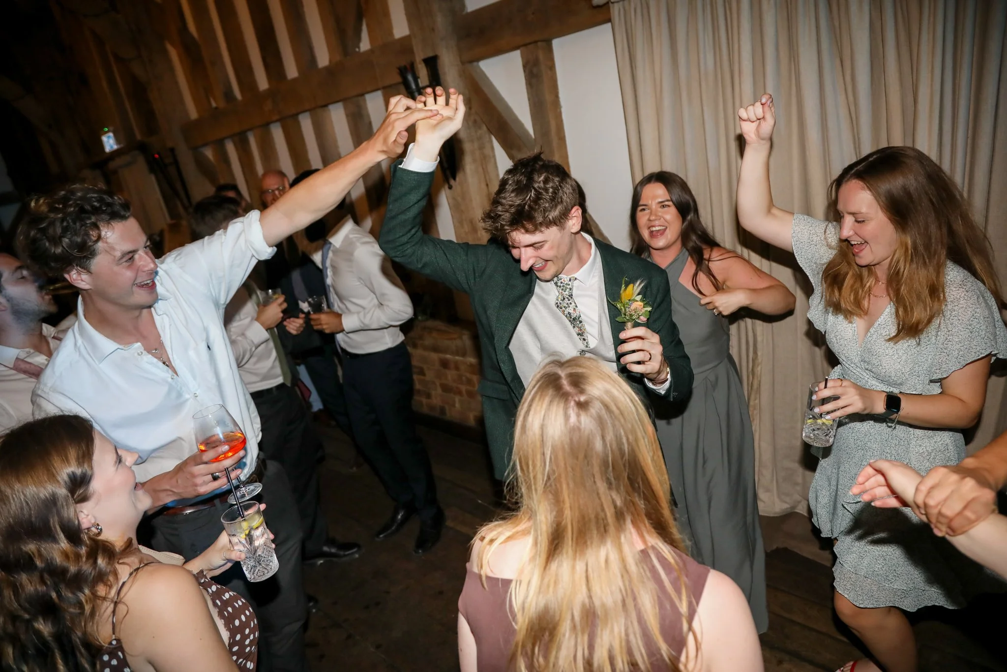 People celebrating at a wedding reception, some with drinks in hand, dancing and smiling in a rustic venue.