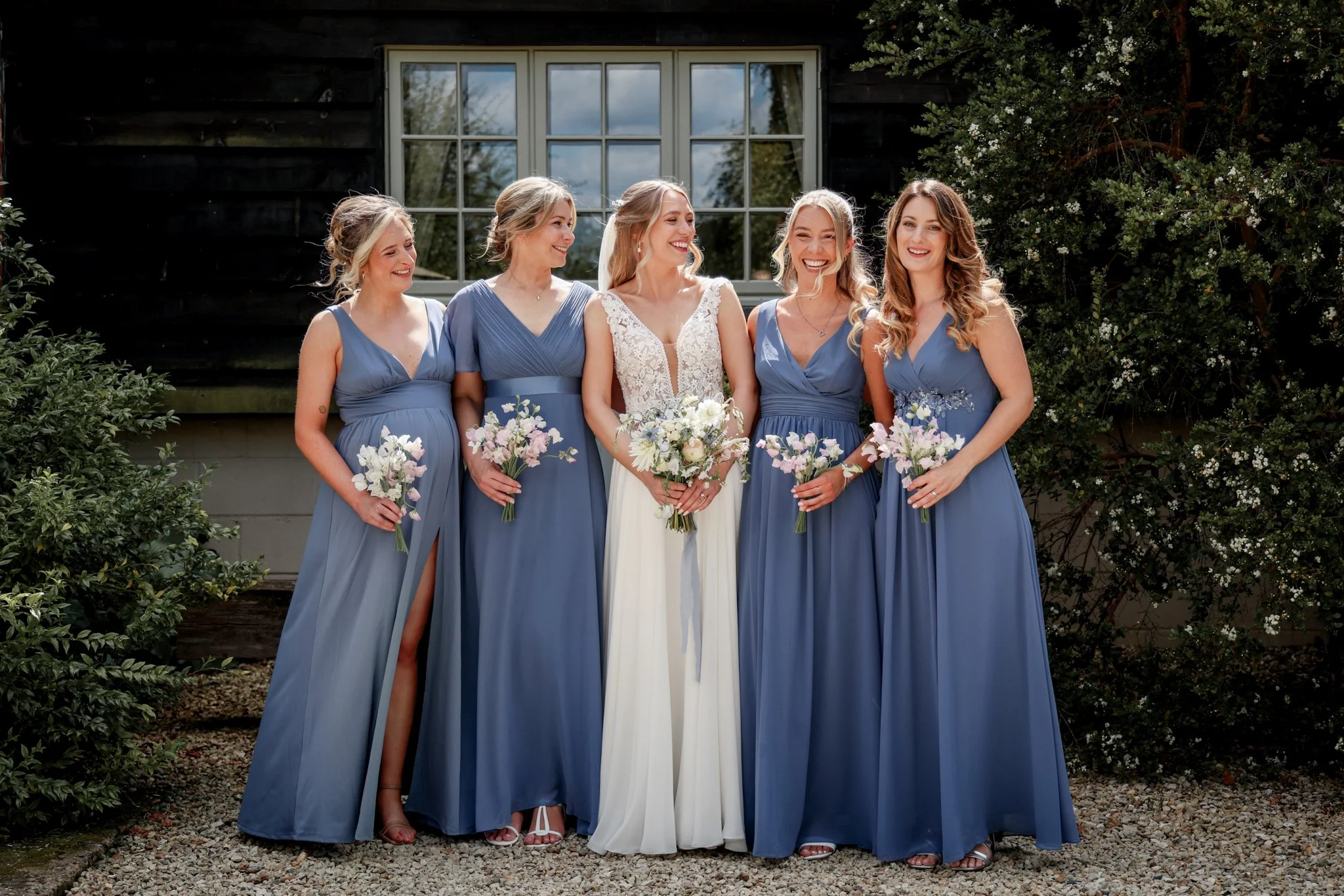 Six women dressed in blue gowns and a bride in a white wedding dress holding bouquets of flowers, standing outdoors in front of a black building with a window, smiling and looking at each other.