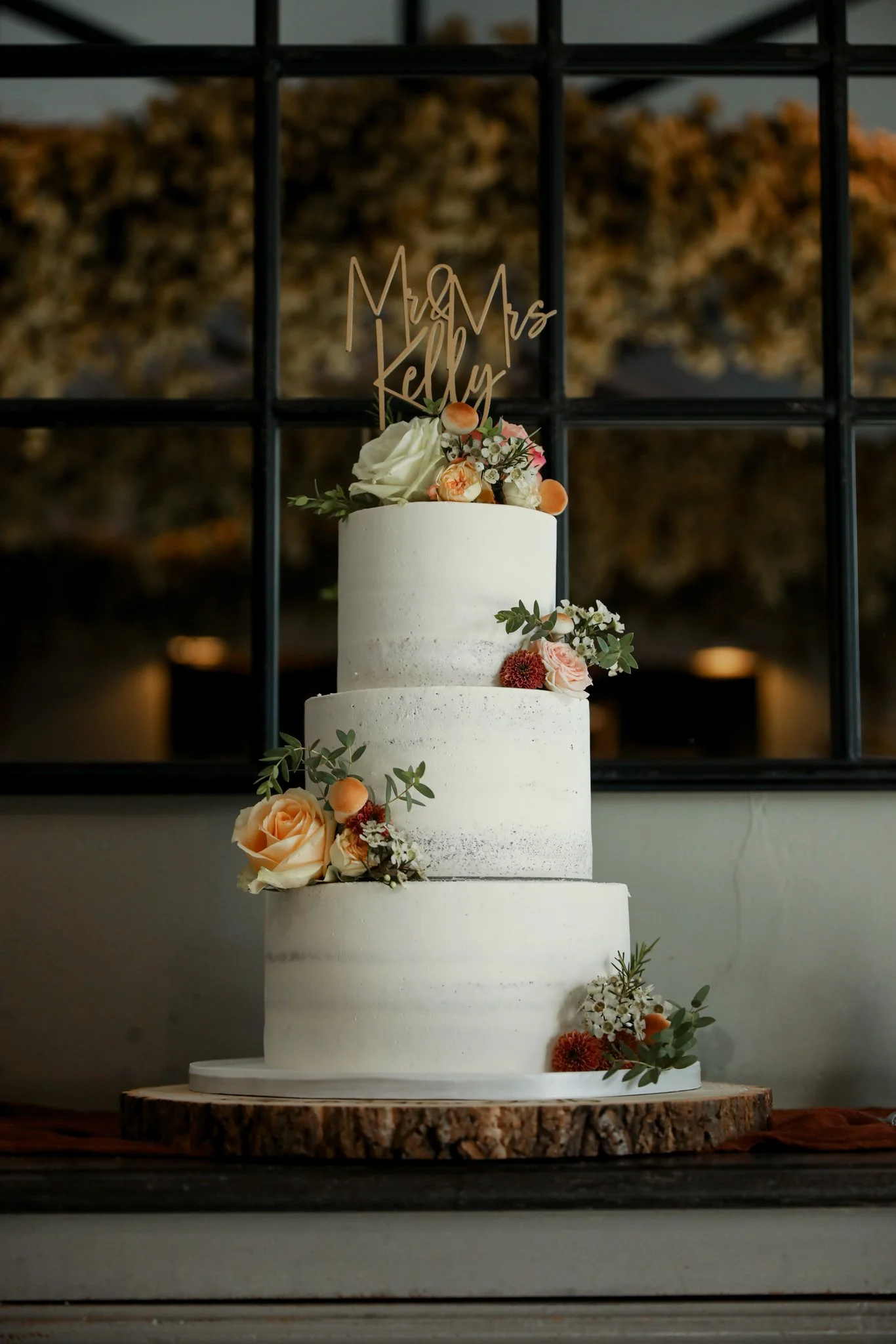 A three-tier white wedding cake decorated with peach, white, and red flowers, topped with a 'Mr & Mrs Kelly' cake topper, placed on a wooden slab against a dark background and black grid window.