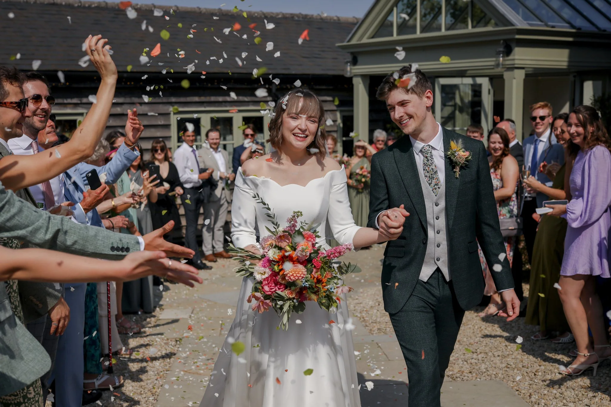 A bride and groom walking hand-in-hand amidst guests celebrating their wedding, with confetti falling around them outside a house.