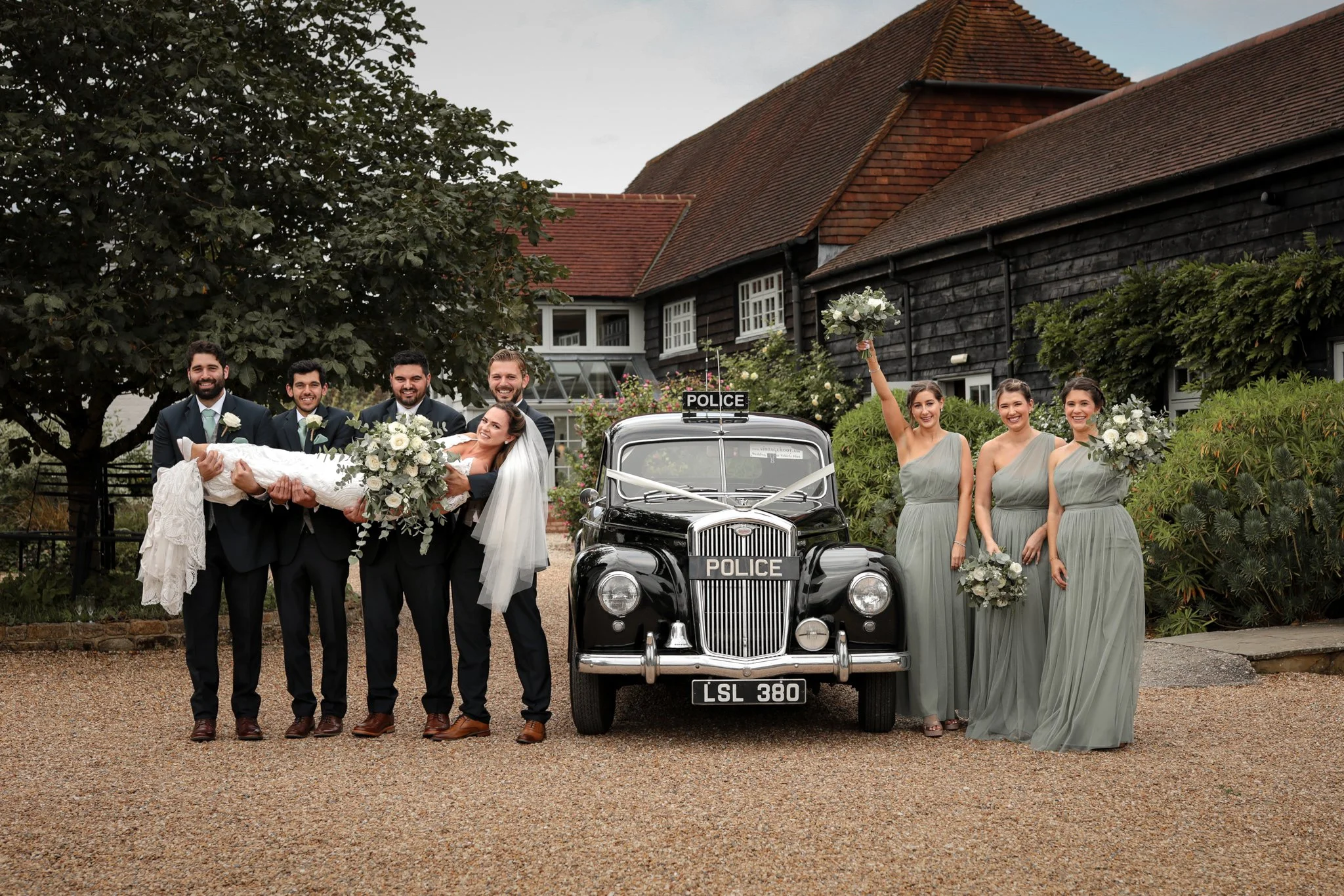 A group of wedding guests, including the bride and groom, pose with a vintage police car outside a house with a dark wooden exterior and a tiled roof. The groom is holding the bride, and a woman in a grey dress is holding a bouquet and raising her ar