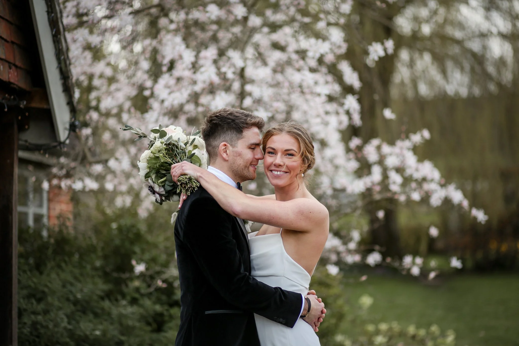 A newlywed couple stands close, embracing outdoors with pink flowering trees in the background. The groom, in a black tuxedo, gently holds the bride, who is smiling and wearing a white wedding dress, holding a bouquet of white flowers and green folia