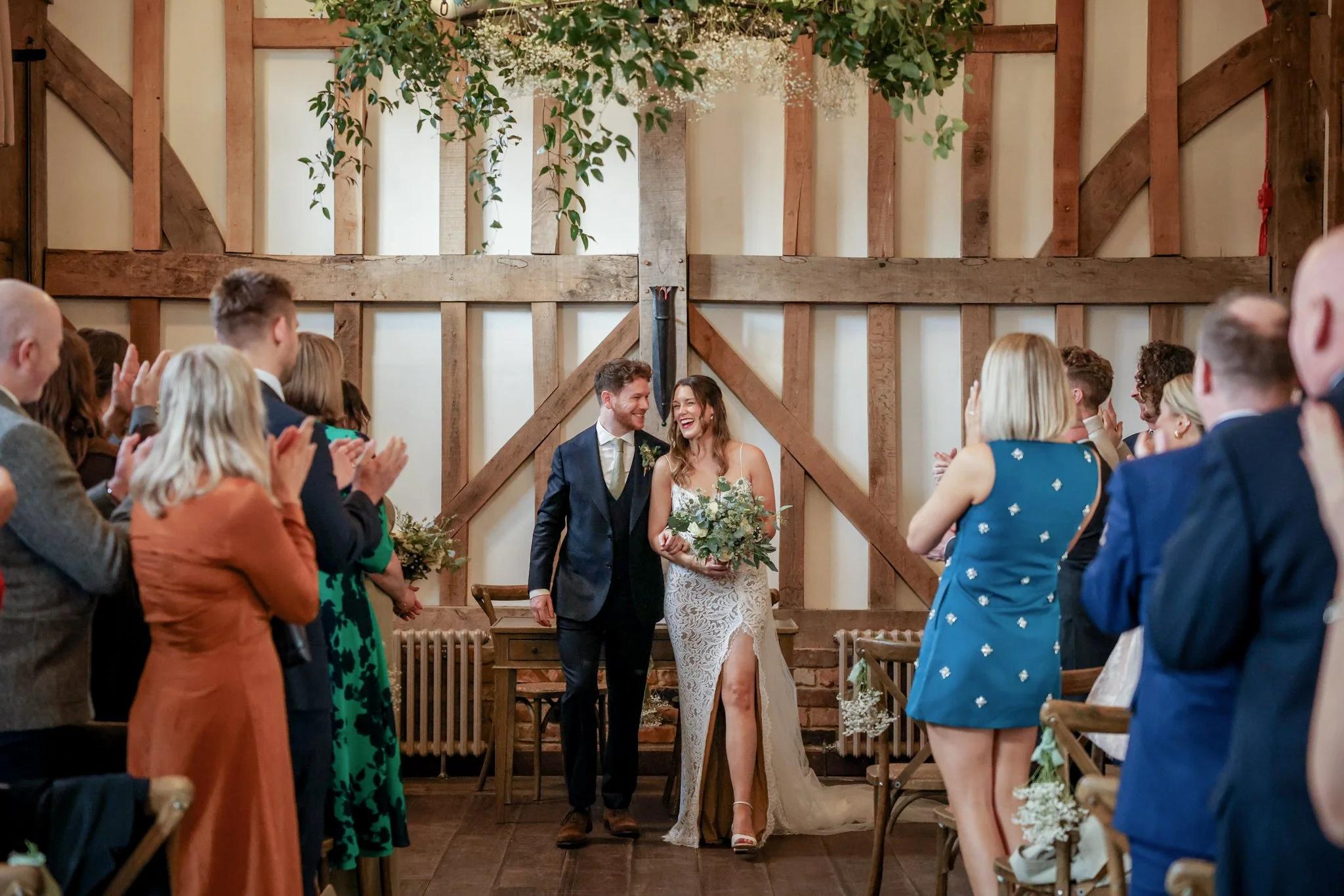 A bride and groom are standing in front of an audience at their wedding ceremony inside a rustic wooden barn, surrounded by friends and family who are clapping and smiling.