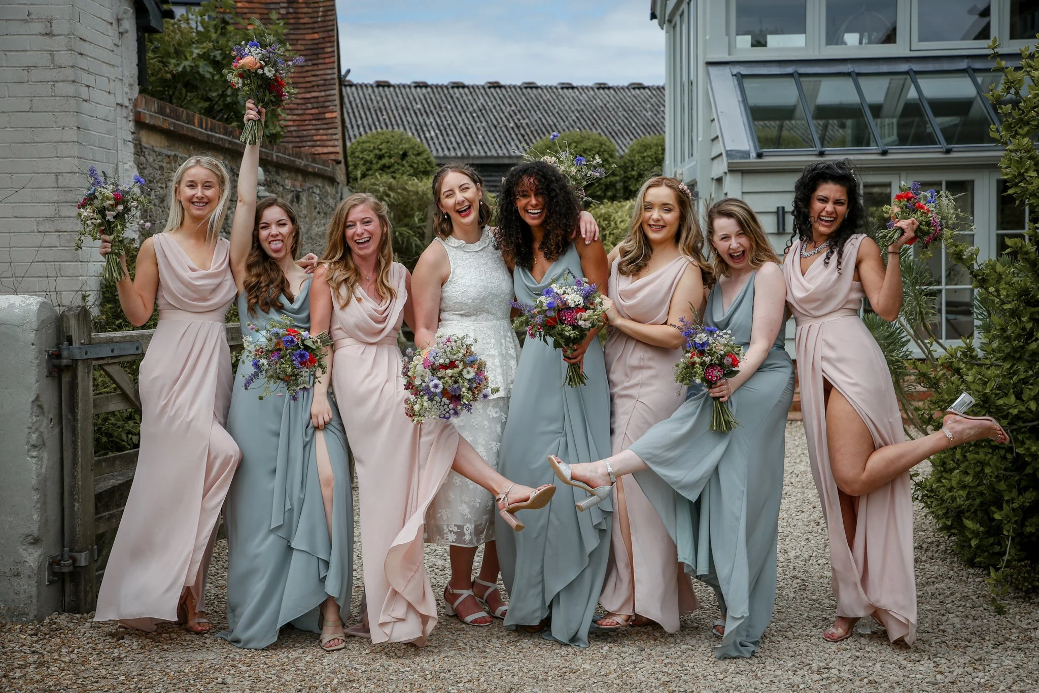 A group of women, some in bridesmaid dresses and one in a wedding dress, smiling and laughing outdoors, holding bouquets of flowers, celebrating at a wedding.