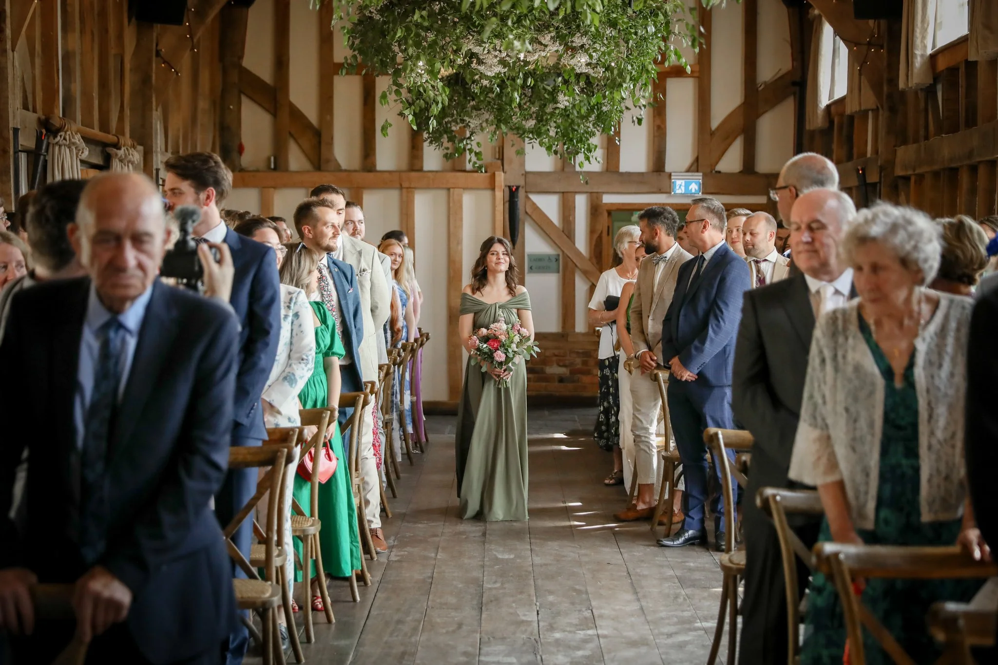 Guests standing in a rustic barn wedding venue with wooden beams, a high ceiling, hanging greenery, and a floral arrangement at the center, attending a wedding ceremony.