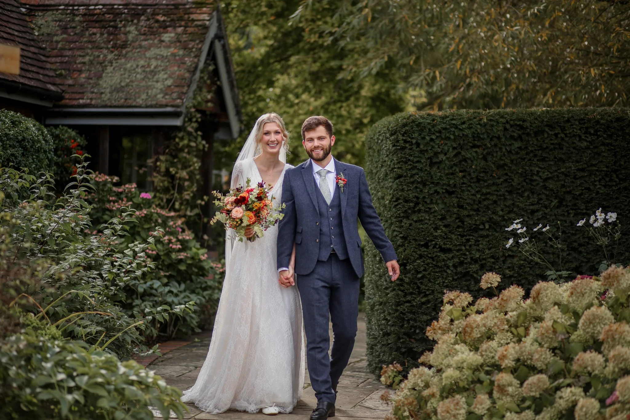 A newlywed couple walking hand in hand on a garden path, surrounded by blooming flowers and greenery, with a rustic house in the background.