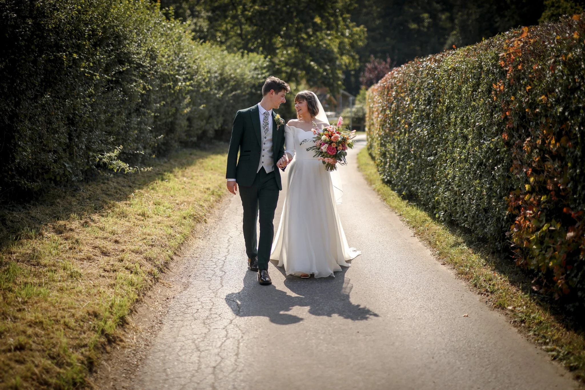 A bride and groom walking hand in hand on a quiet path, surrounded by green and reddish bushes, during daytime. The bride is holding a bouquet of flowers and is wearing a white wedding dress. The groom is in a dark suit with a boutonniere.