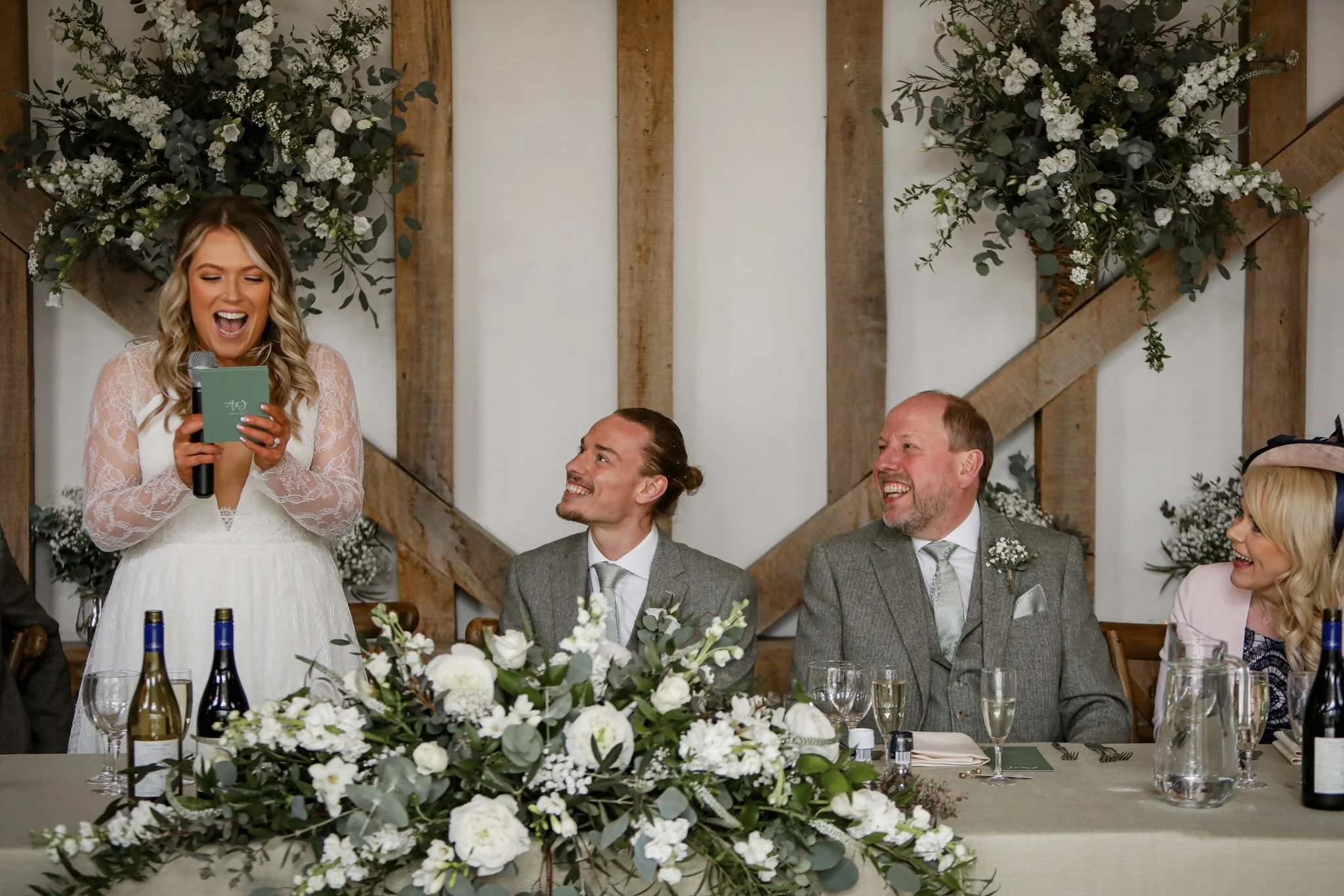 A woman in a white dress giving a speech at a wedding reception, surrounded by men in suits sitting at a table decorated with flowers, wine bottles, and glasses, against a backdrop with floral arrangements and wooden accents.
