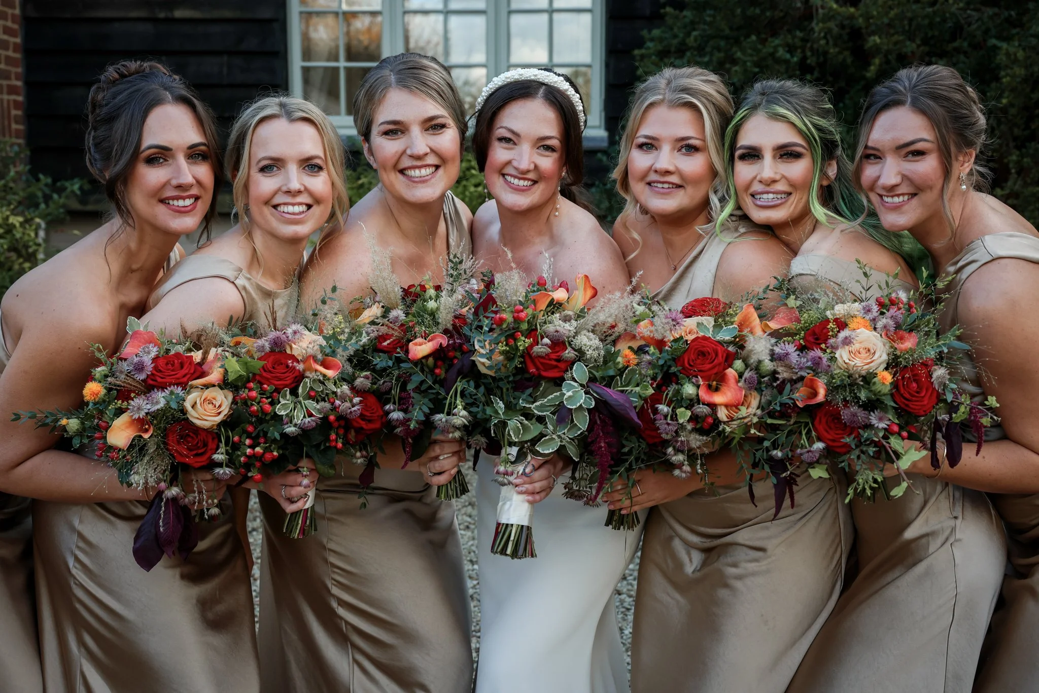Group of seven smiling women in beige dresses holding bouquets of colorful flowers, standing outdoors in front of a black building and greenery.