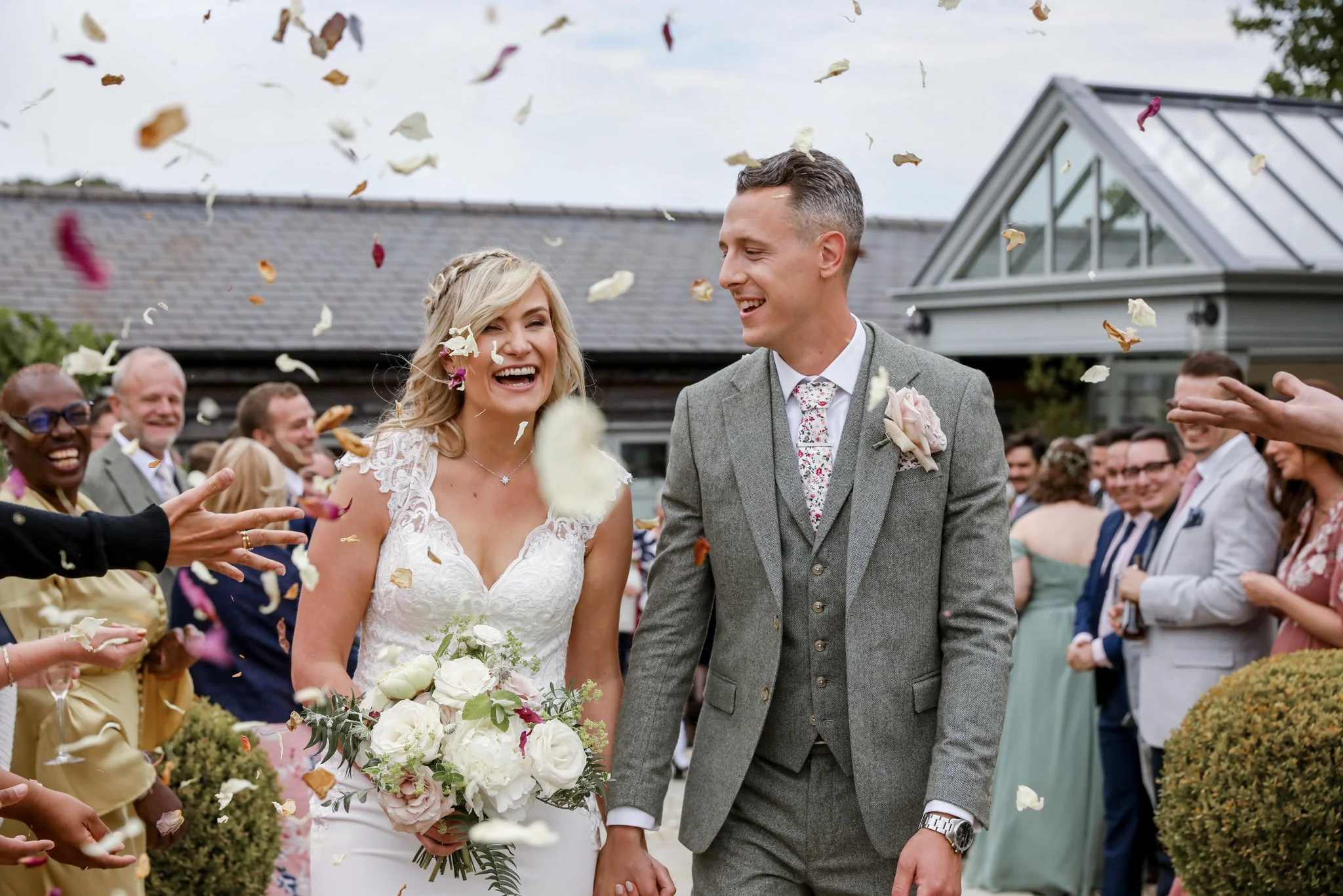 A bride and groom smiling and holding hands during their outdoor wedding celebration, surrounded by guests throwing flower petals.