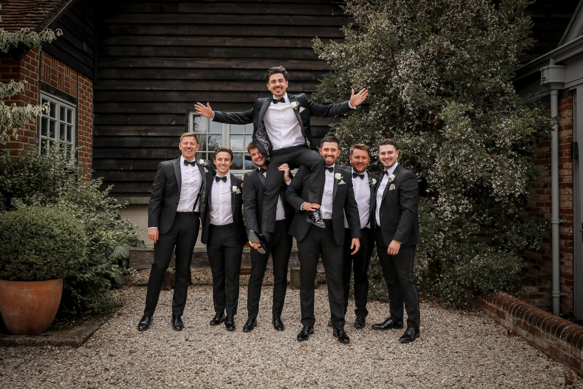 A group of seven men in formal tuxedos, with one man lifted onto the shoulders of two others, celebrating outdoors in front of a dark wooden wall and a large flowering tree.