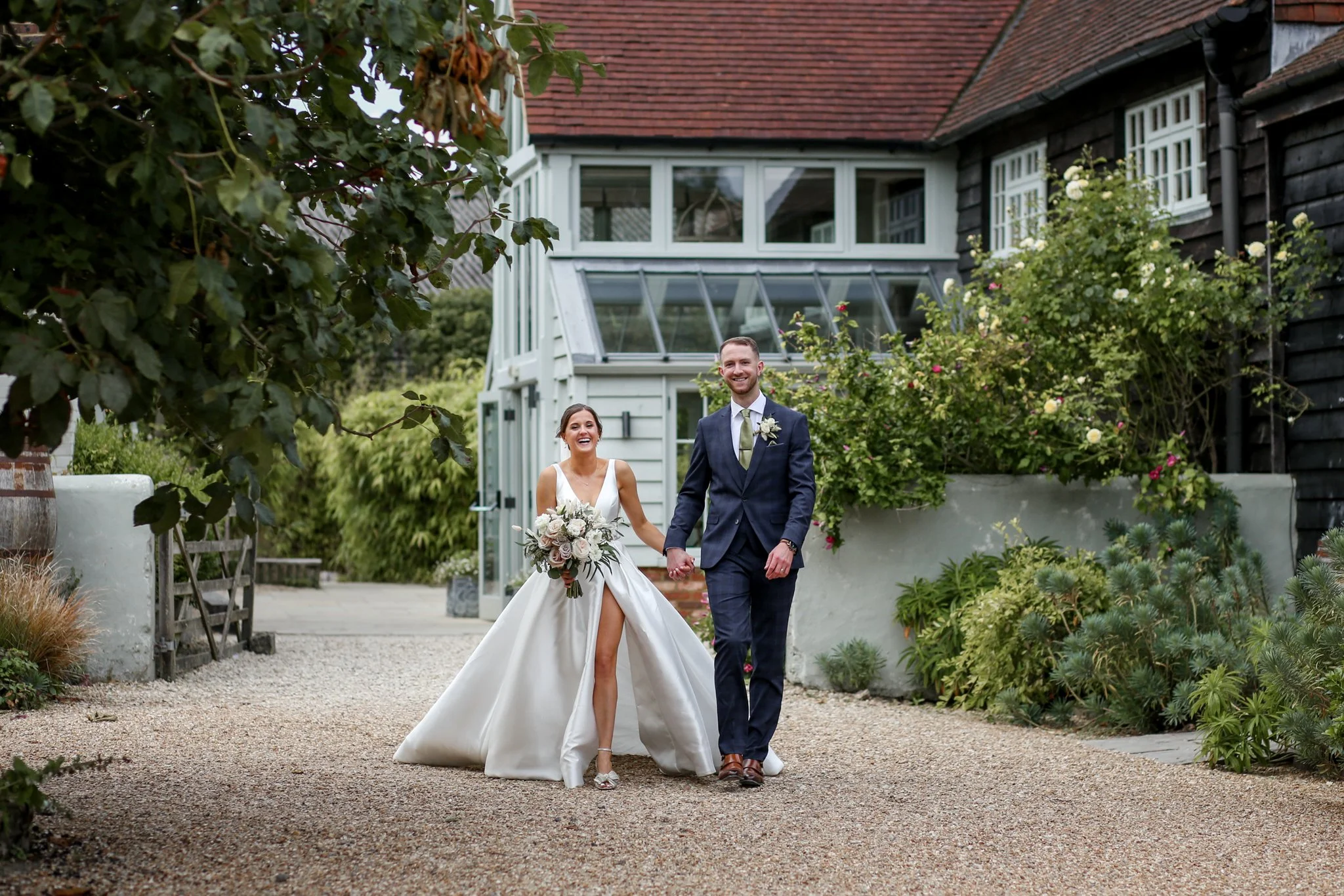 A newlywed couple walking hand in hand outdoors in a garden area, smiling and dressed in wedding attire, with the bride in a white gown holding a bouquet of flowers and the groom in a dark suit with a tie, surrounded by greenery and a rustic house in