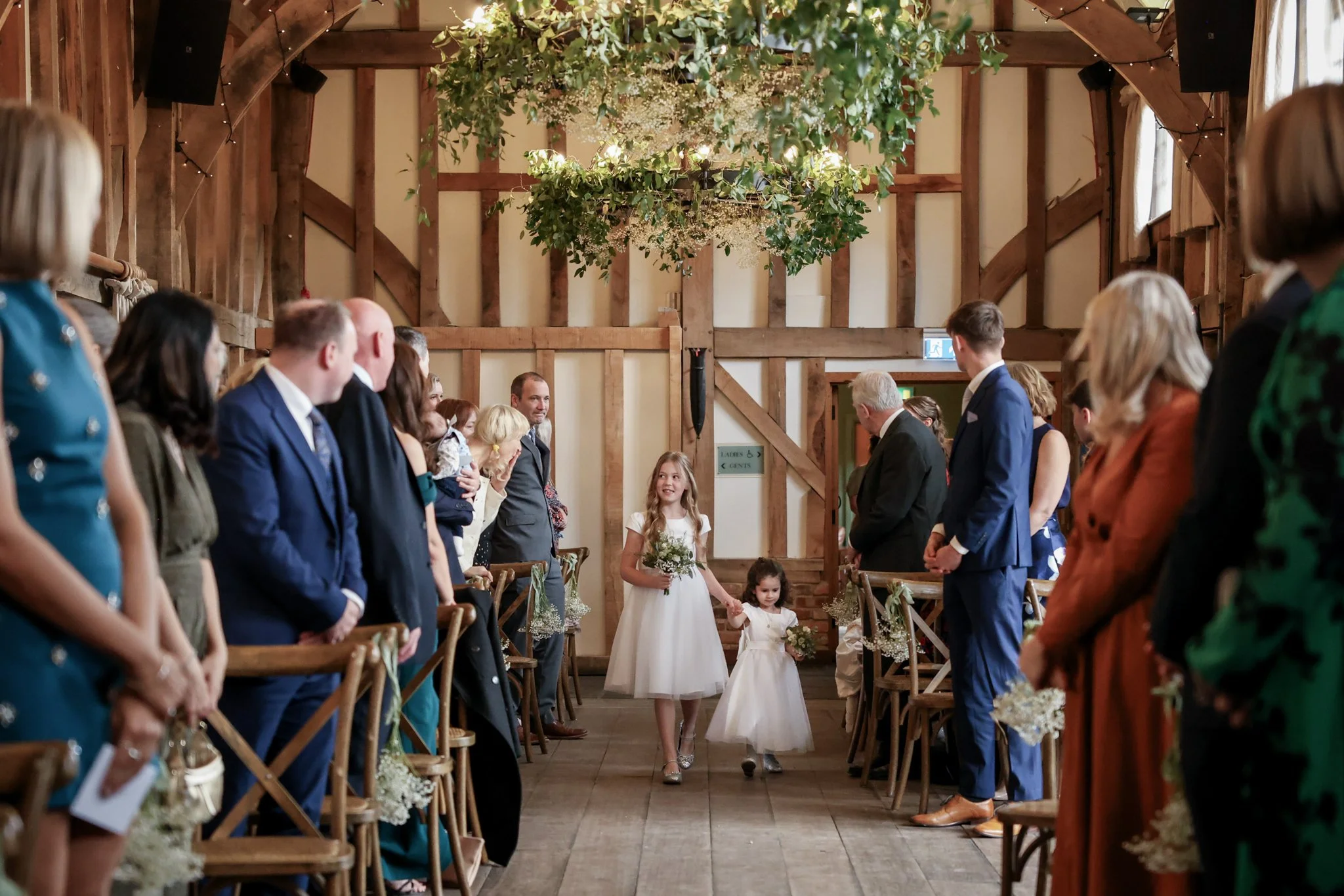 A wedding ceremony inside a rustic barn with wooden beams, decorated with lush greenery and hanging floral arrangements. Guests are seated on wooden chairs, watching two flower girls walking down the aisle.