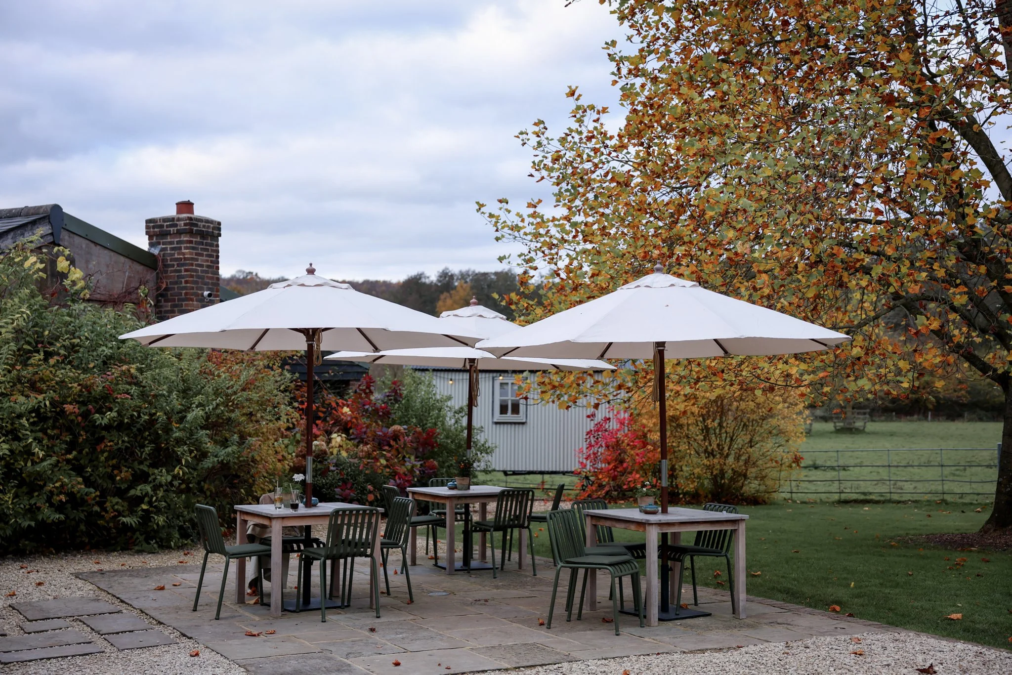 An outdoor patio with several tables and chairs under white umbrellas, surrounded by autumn trees with orange and green leaves, in a garden setting.