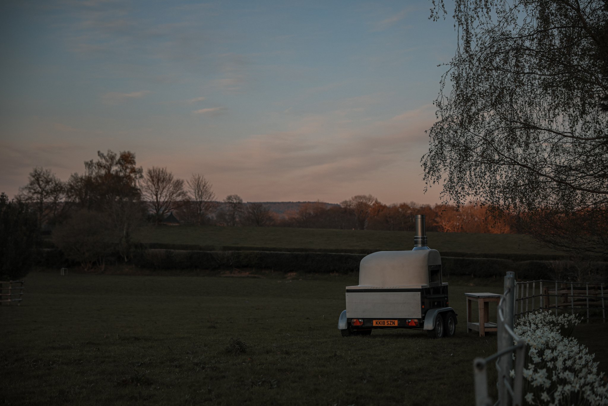 A backyard scene at dusk with a propane heater, a trailer, and trees with autumn foliage in the background.