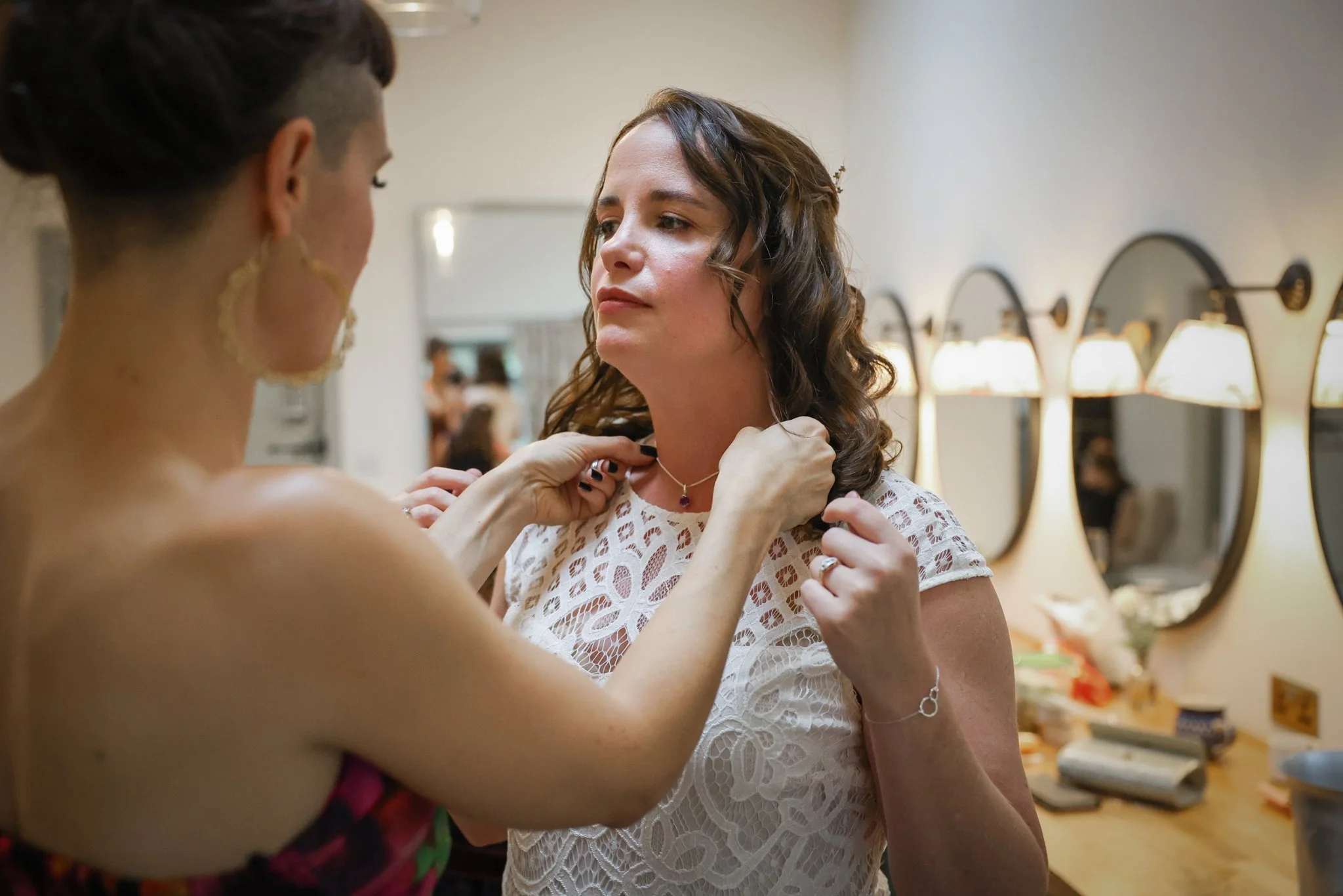 A woman with brown, curly hair in a white lace dress fitting a necklace on another woman with dark hair in an updo and hoop earrings in a room with mirrors and vanity lights.