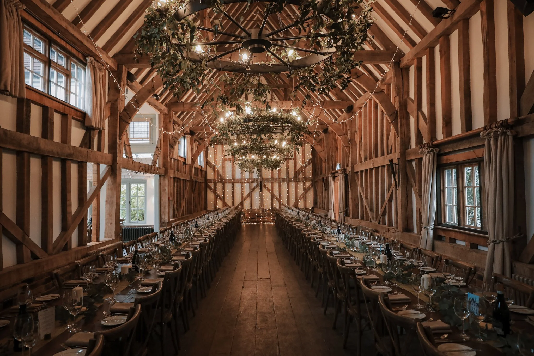 Long banquet table set for a formal event in a rustic wooden barn with ceiling fans, fairy lights, and greenery decorations.