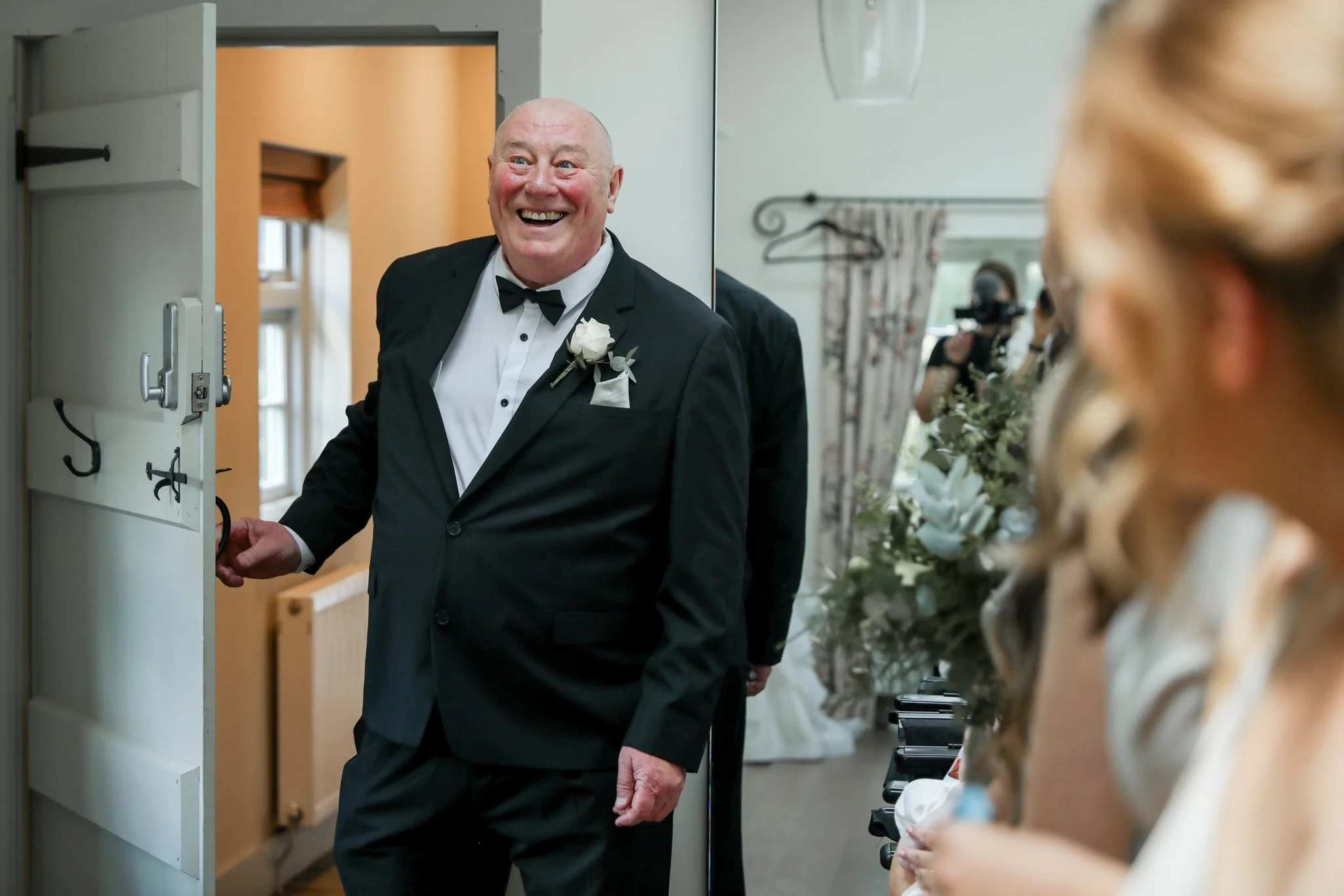A cheerful man in a tuxedo with a white corsage on his lapel is smiling and greeting guests at a wedding reception, reflected in a mirror.
