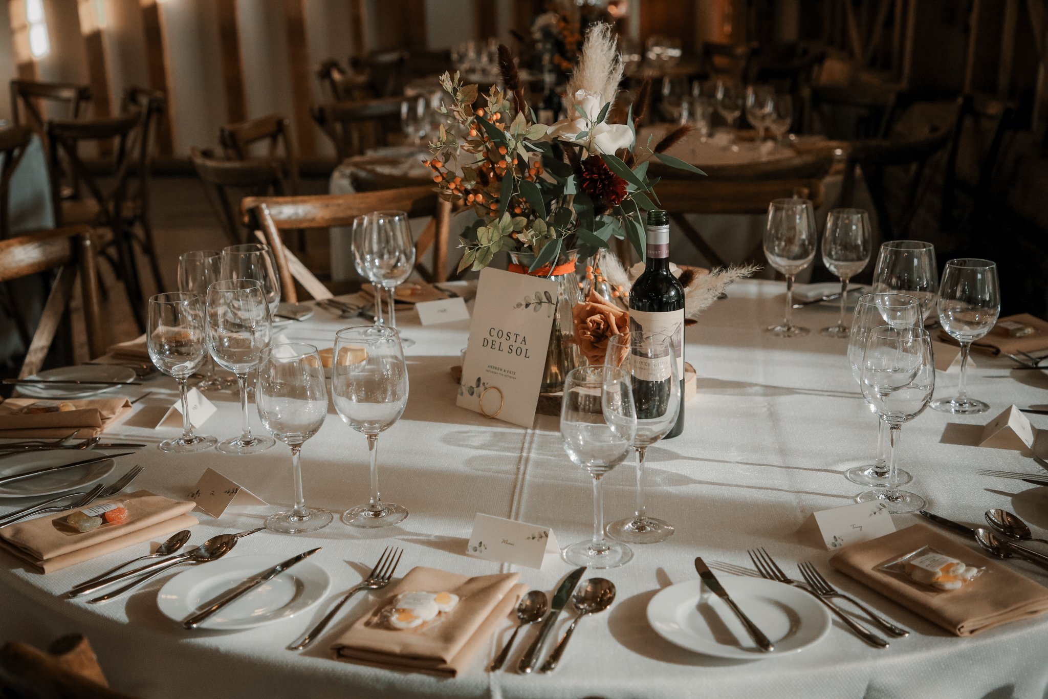 Elegant banquet table setup with wine glasses, silverware, napkins, and a floral centerpiece with greenery, white and red flowers, and pampas grass in a rustic wooden venue.