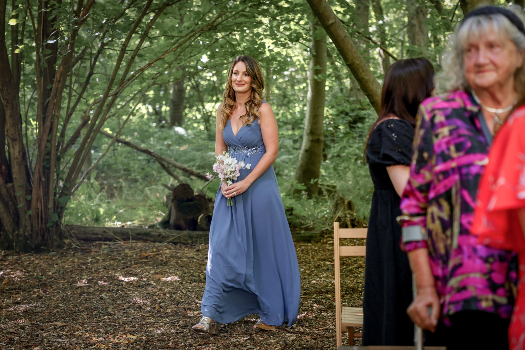 A woman in a blue dress holding a bouquet standing in a wooded area during the daytime, with women in colorful clothing partially visible on the right side.