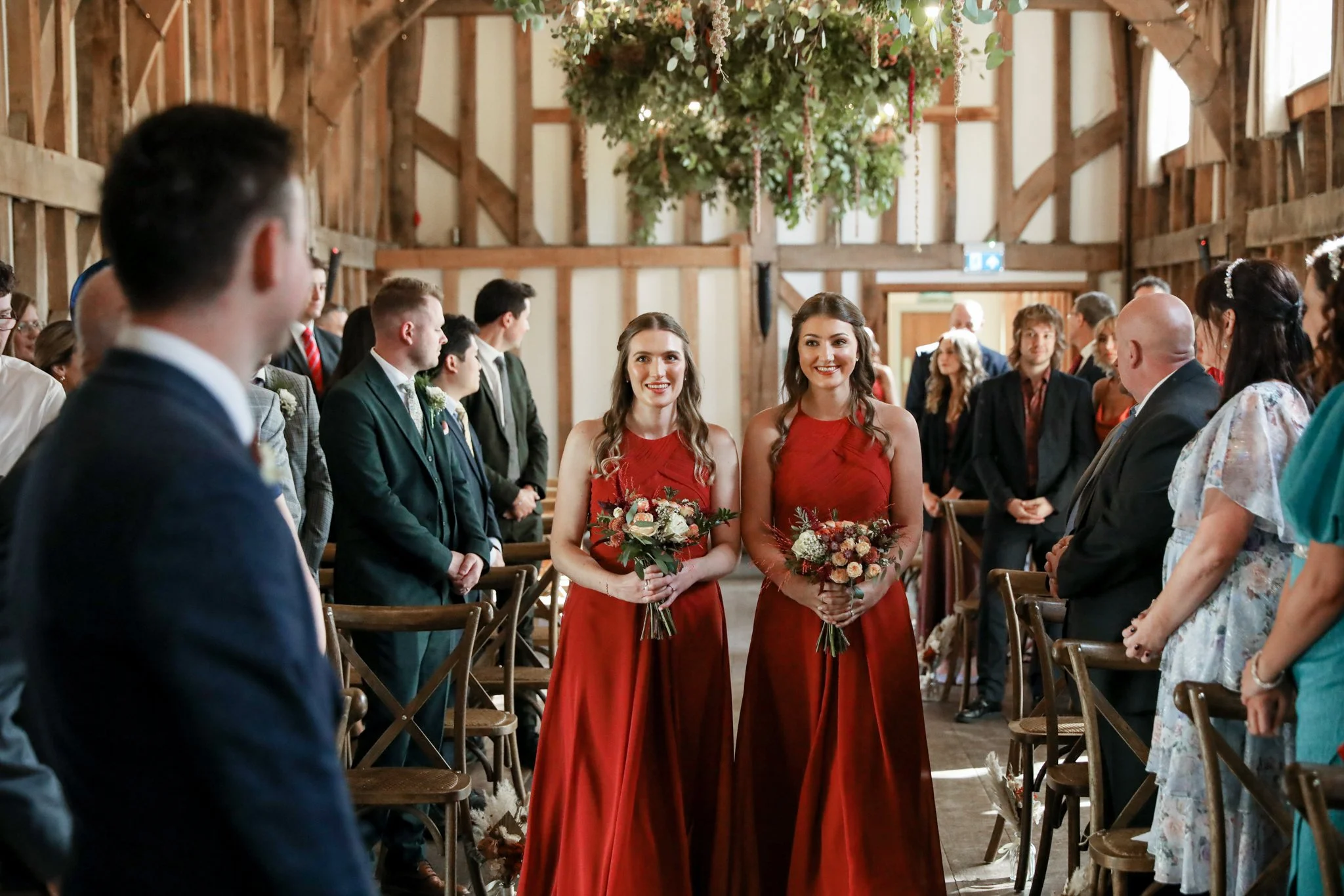 Two women in matching red dresses holding bouquets of flowers, standing in a rustic wedding ceremony inside a barn with wooden beams and hanging floral decorations, surrounded by seated guests.