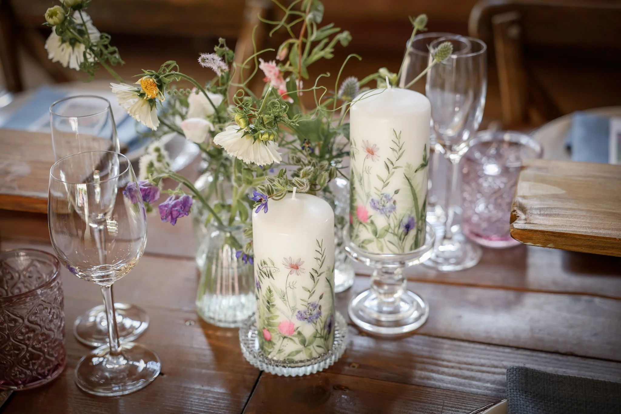 A table centerpiece with a bouquet of mixed flowers, including white, purple, and pink blossoms, in clear glass vases, flanked by white candles, on a wooden table surrounded by empty wine glasses.