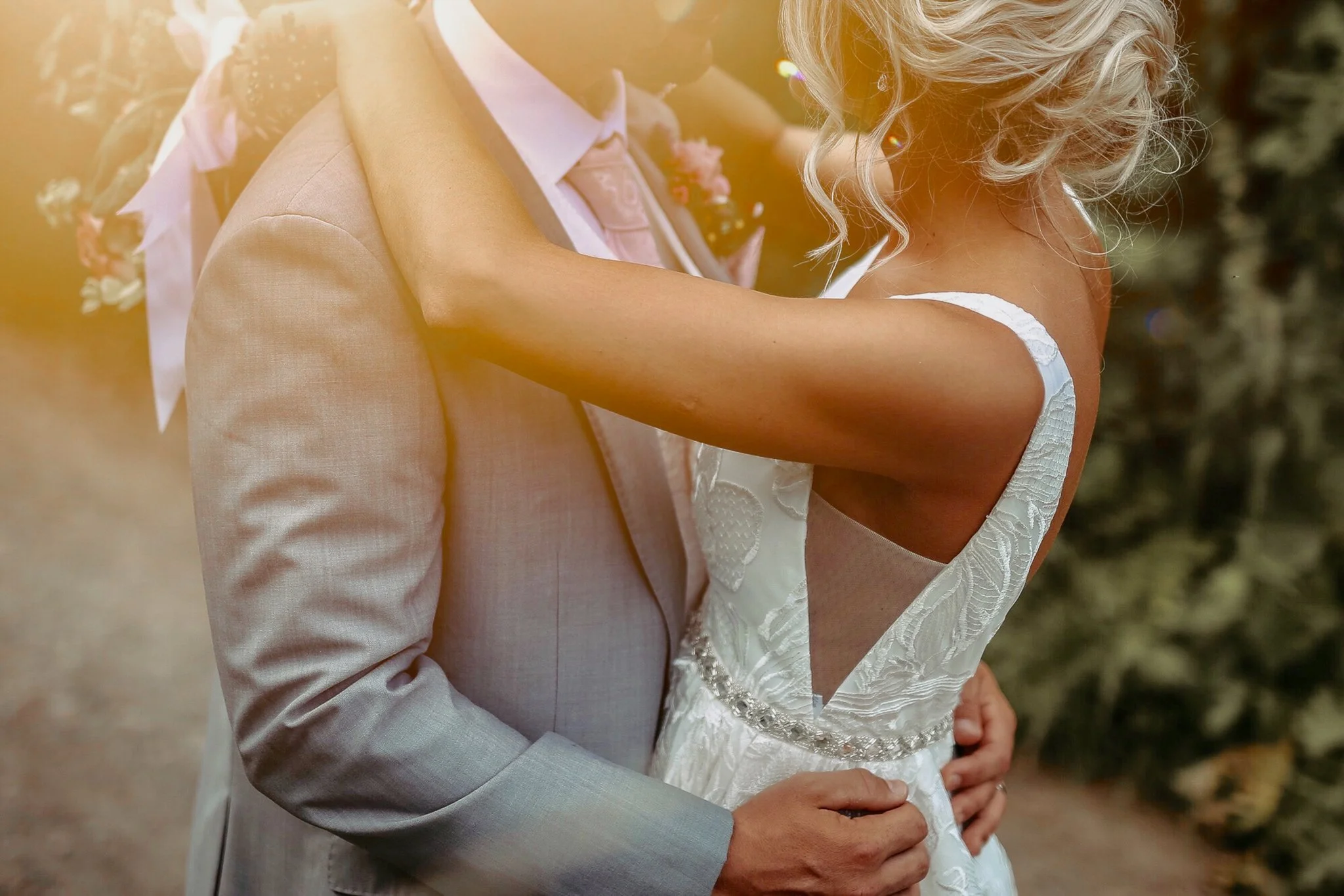 Close-up of a bride and groom embracing outdoors, holding each other lovingly, with warm sunlight in the background.