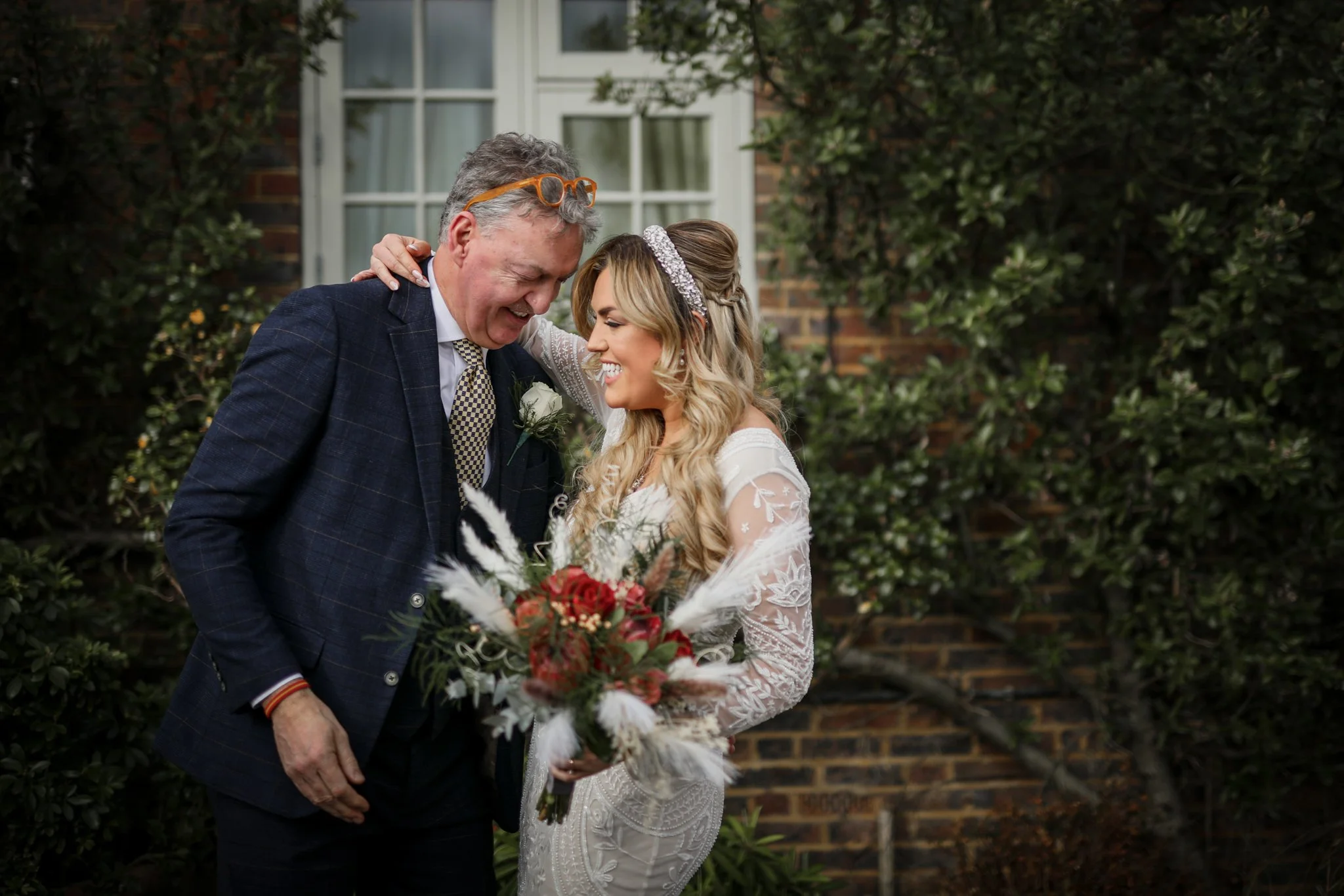 A bride and an older man, possibly her father, hugging and smiling at each other during a wedding ceremony outdoors. The bride is holding a bouquet of flowers.