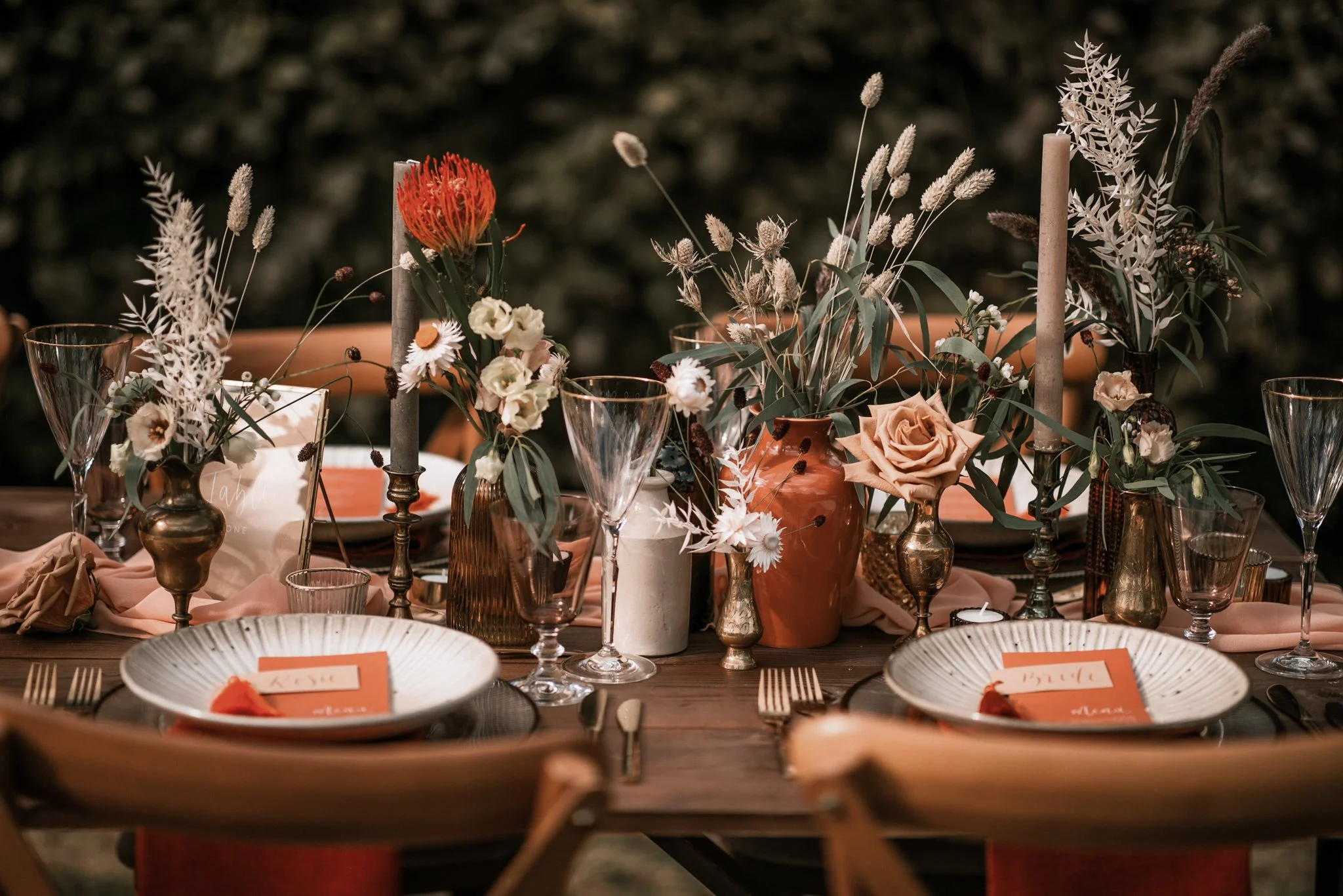 A decorated table with various vases containing flowers and greenery, candles, and place settings with orange napkins and name cards, set for an event or celebration.
