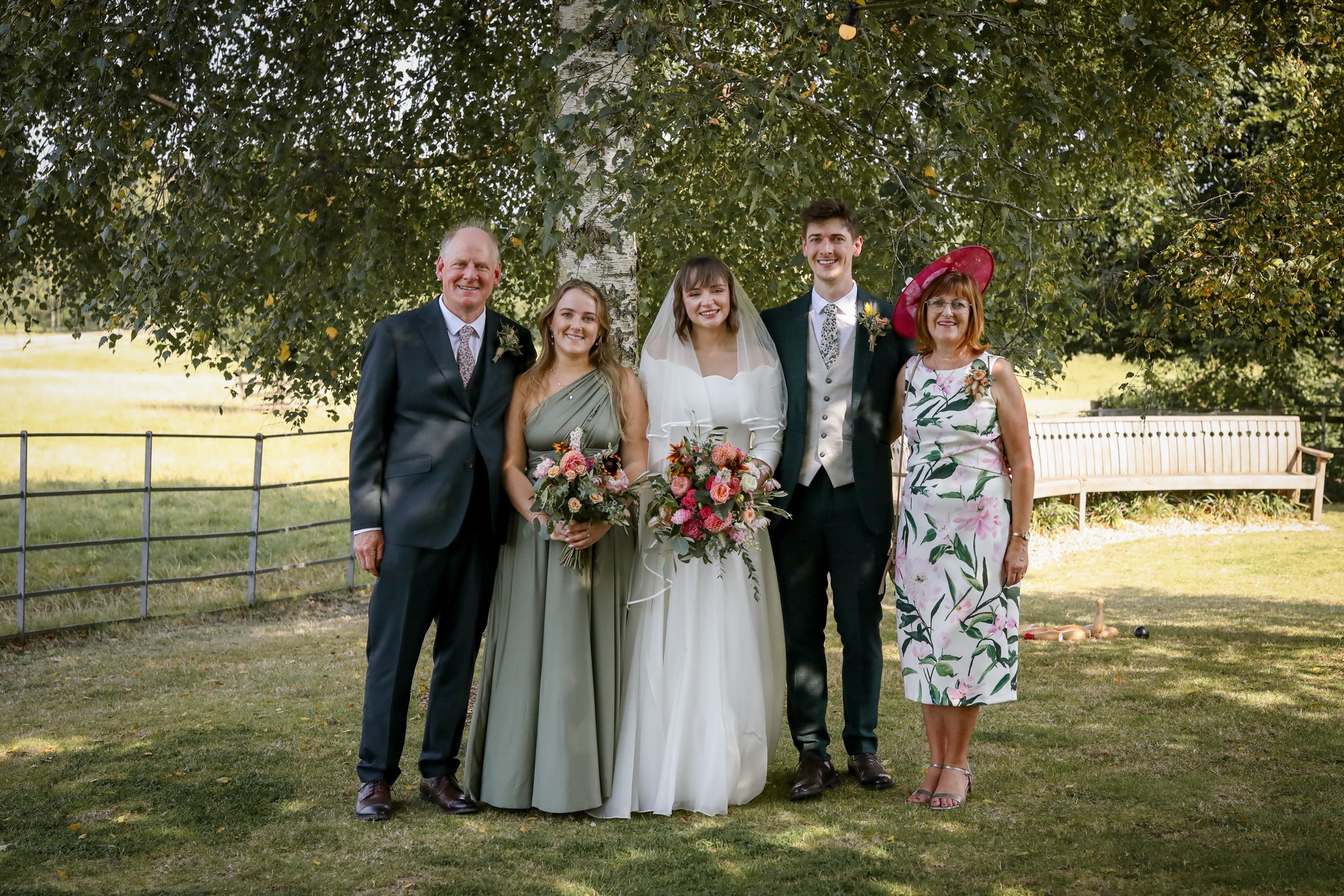 A group of five people dressed in formal attire standing outdoors under a large tree, with a bride and groom in the center holding bouquets, smiling for a photo during a wedding.
