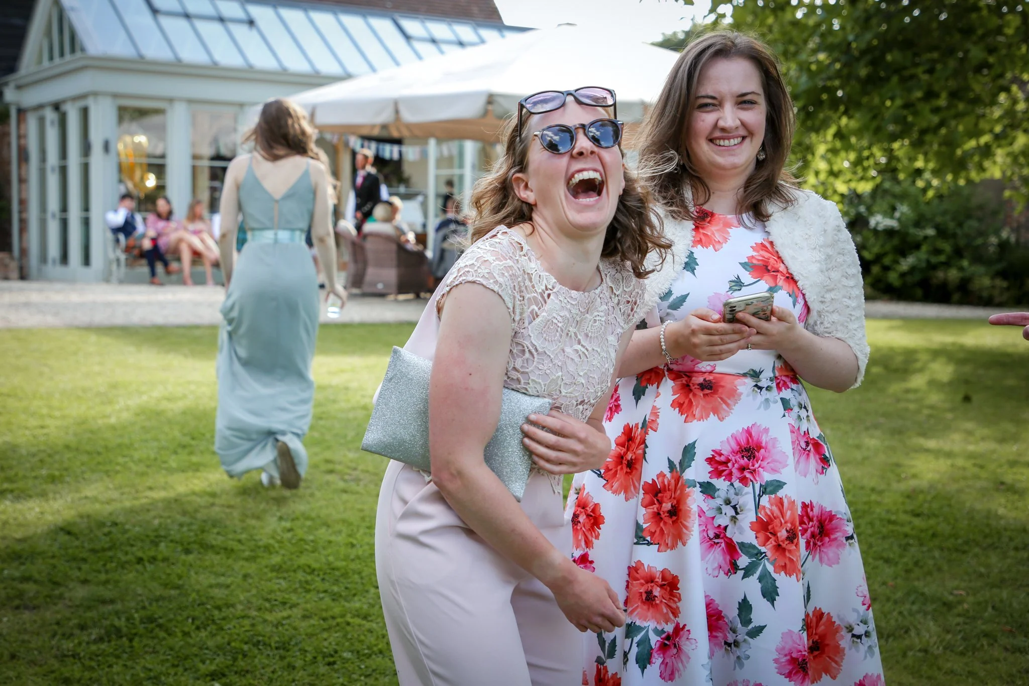 Two women smiling and laughing at an outdoor gathering, with one holding a phone and the other carrying a silver purse. In the background, other guests are socializing near a house with a large white umbrella.