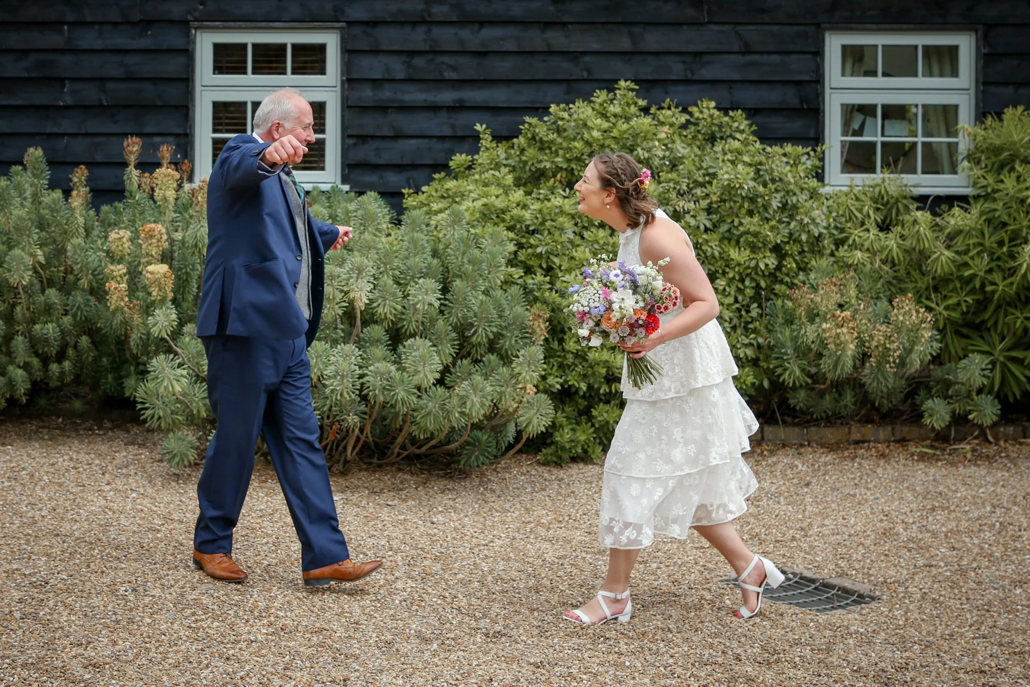A man and woman dressed in wedding attire are outside, smiling and interacting. The woman is holding a bouquet and wearing a white dress, while the man is wearing a dark blue suit. They appear to be happy and engaged in a joyful moment.